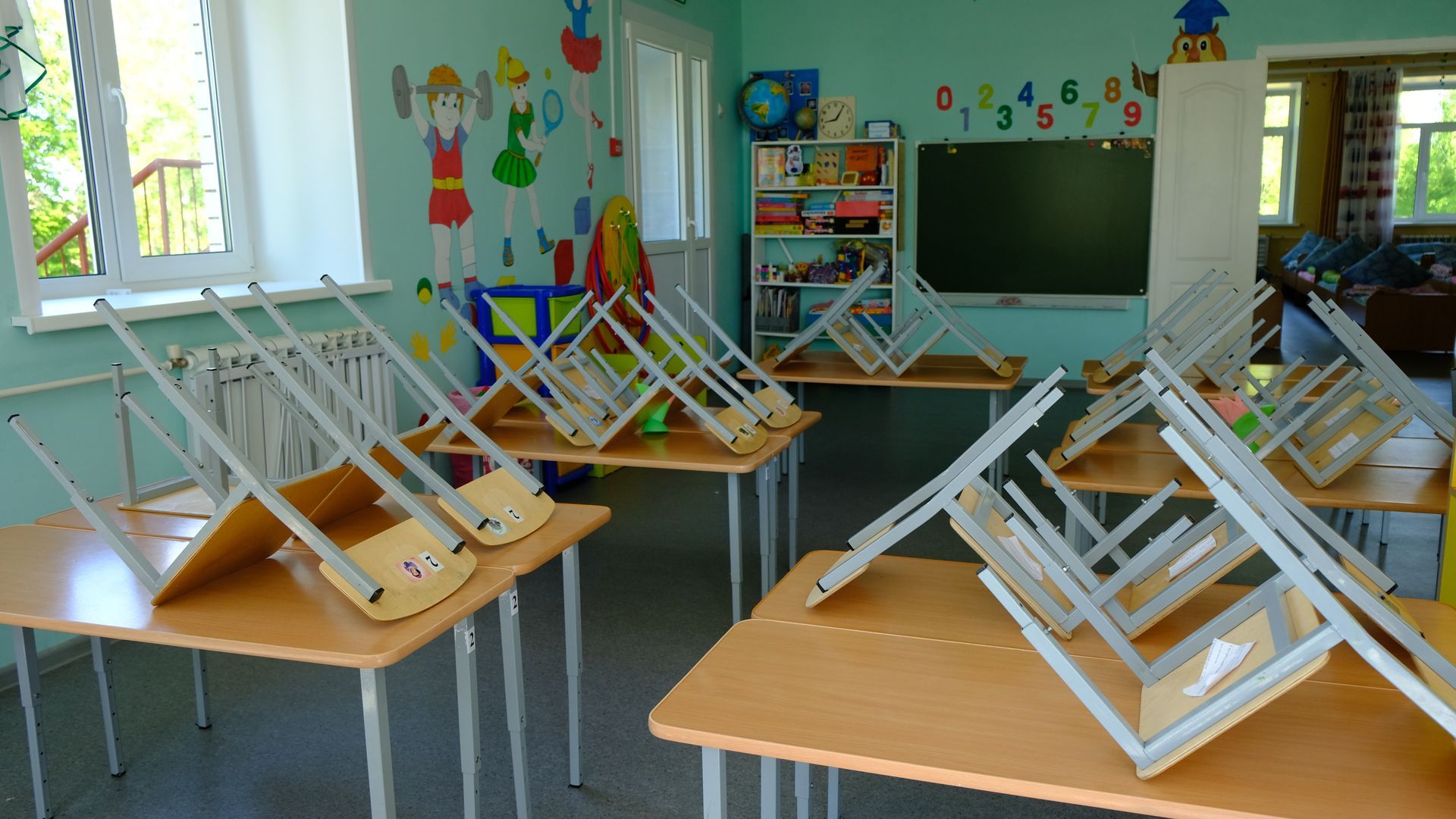 A classroom with light green walls features several wooden desks with chairs stacked upside down on top of them.