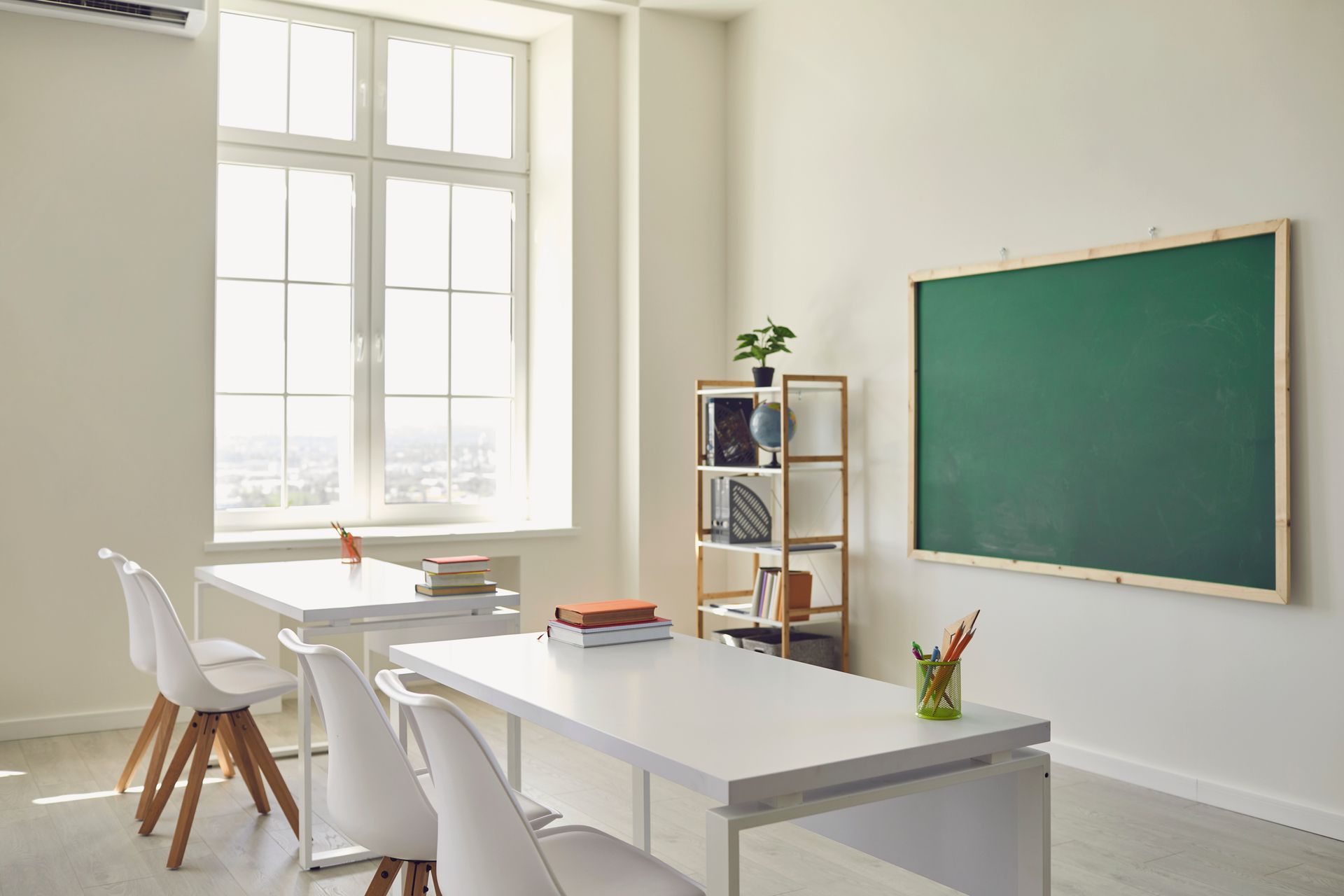 A bright, minimalist classroom with white tables, chairs, a bookshelf, and a blank green chalkboard on a white wall.