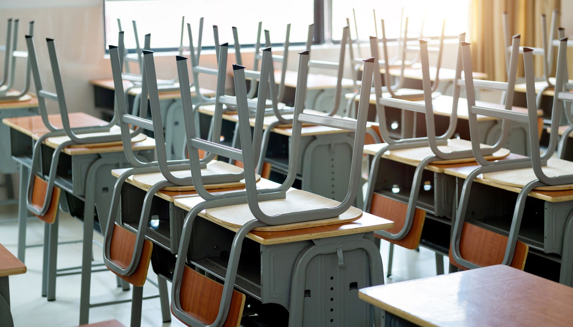 A classroom featuring rows of wooden desks with metal frames, each topped with an inverted chair.