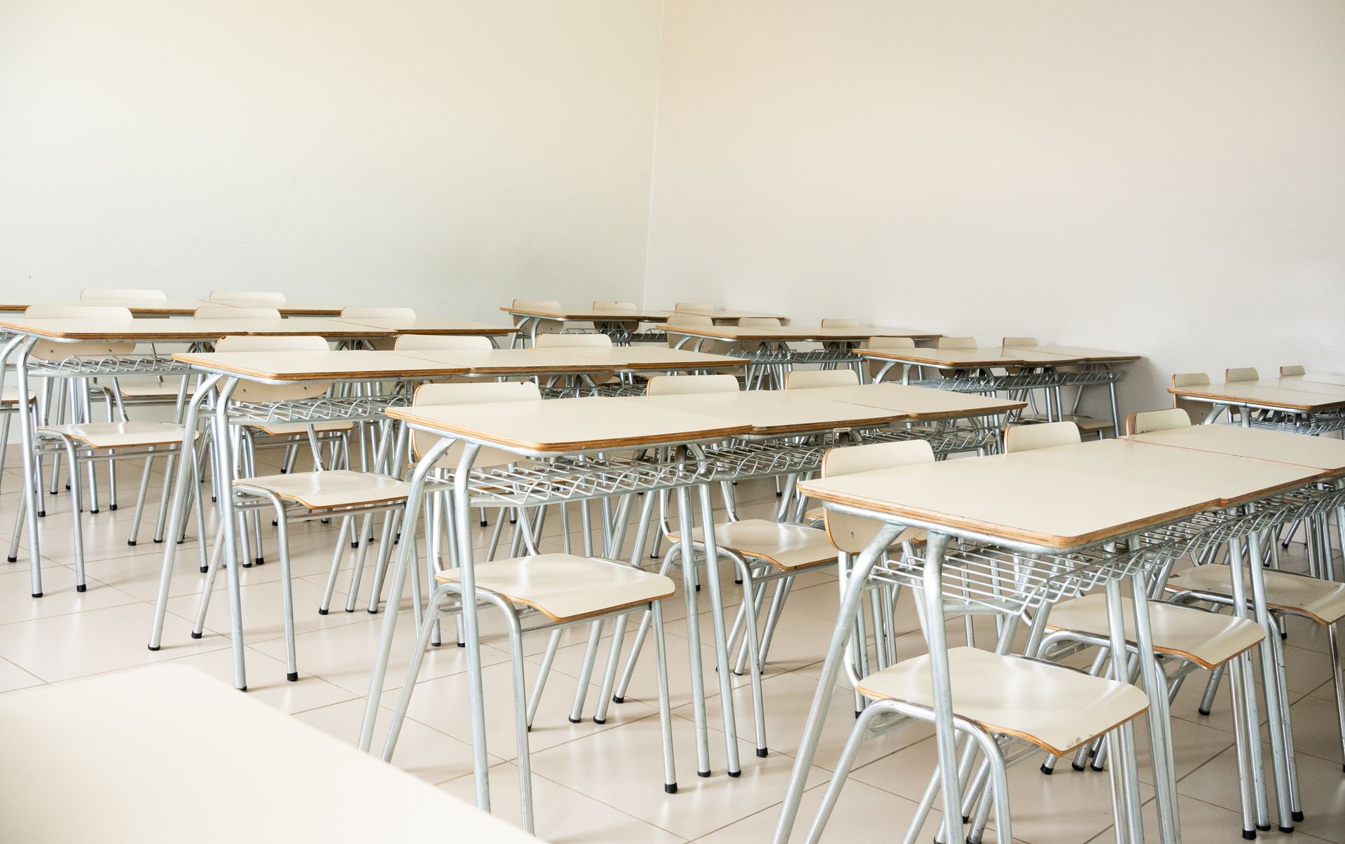 A classroom featuring rows of wooden desks with metal frames and matching chairs set against plain, off-white walls.