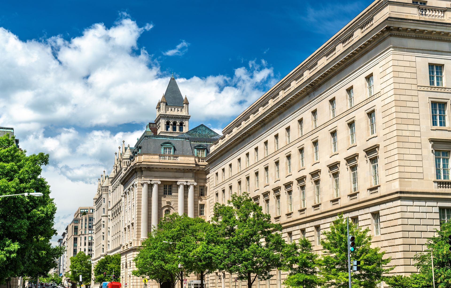 A tall, ornate beige building with a dark-roofed tower set against a bright blue sky with white clouds and green trees.