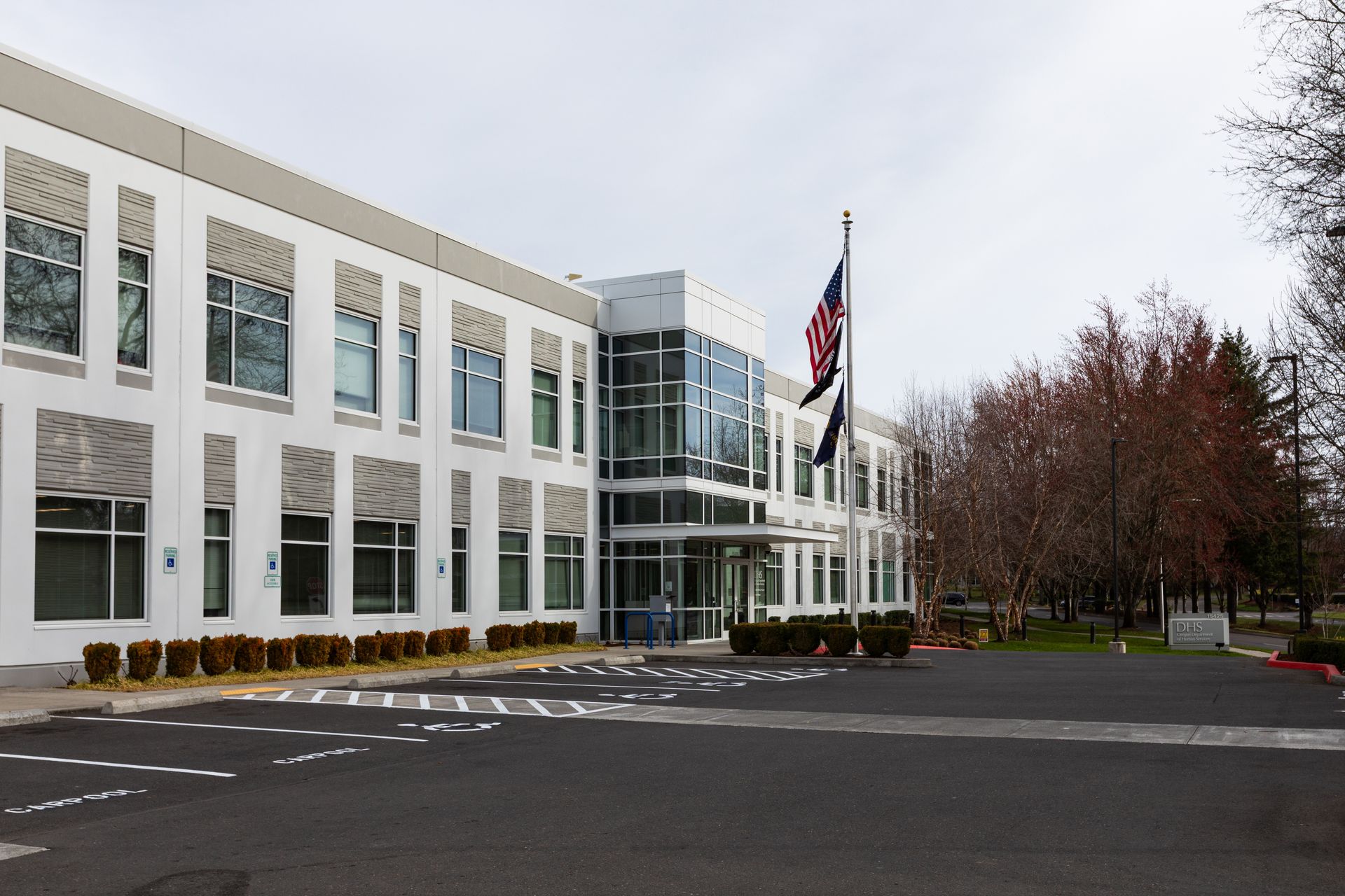 A modern, two-story white office building with large windows and a glass entryway flying an American flag in a parking lot.