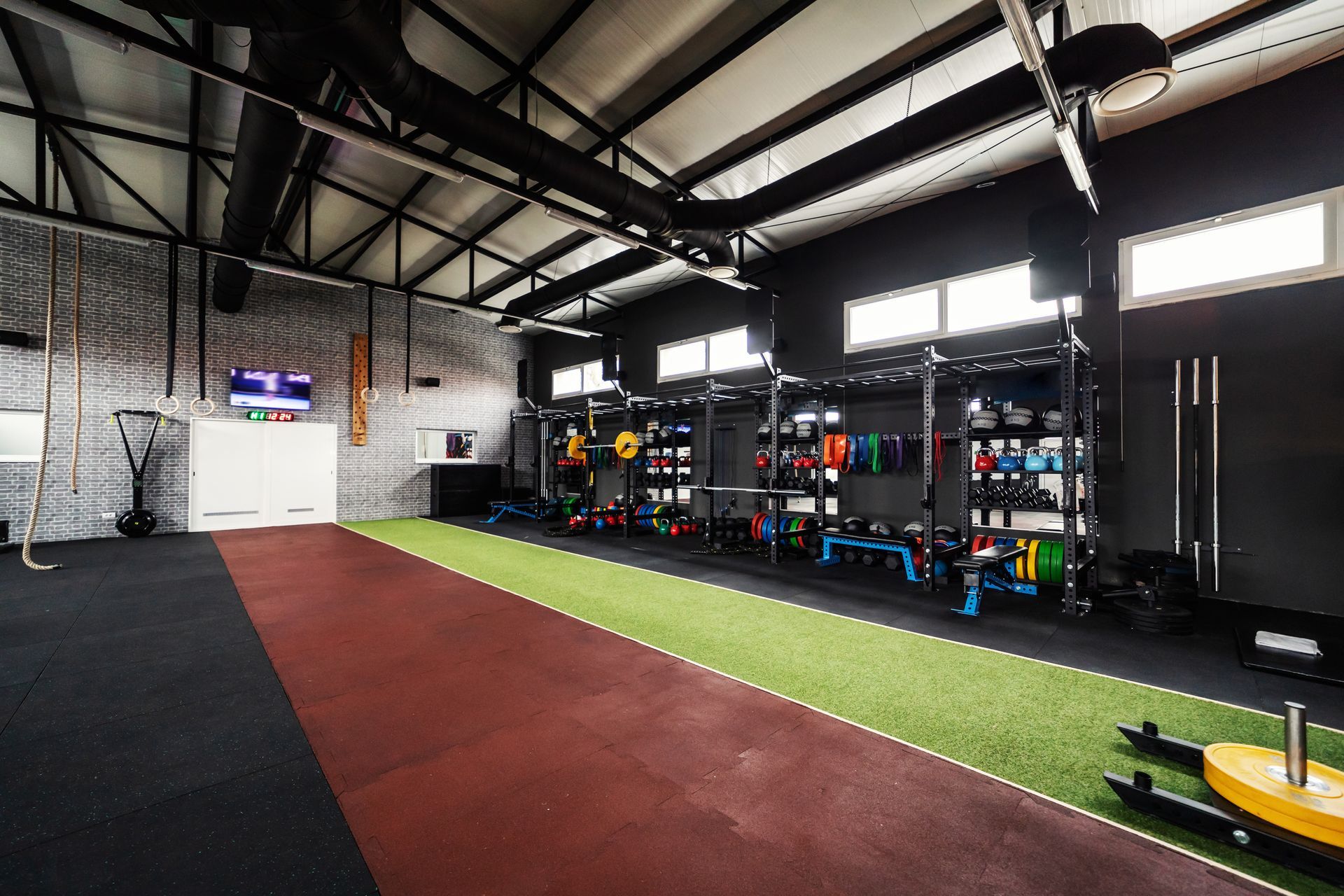 Modern gym interior featuring a green turf lane, rubber flooring, fitness equipment storage, and high industrial ceilings.