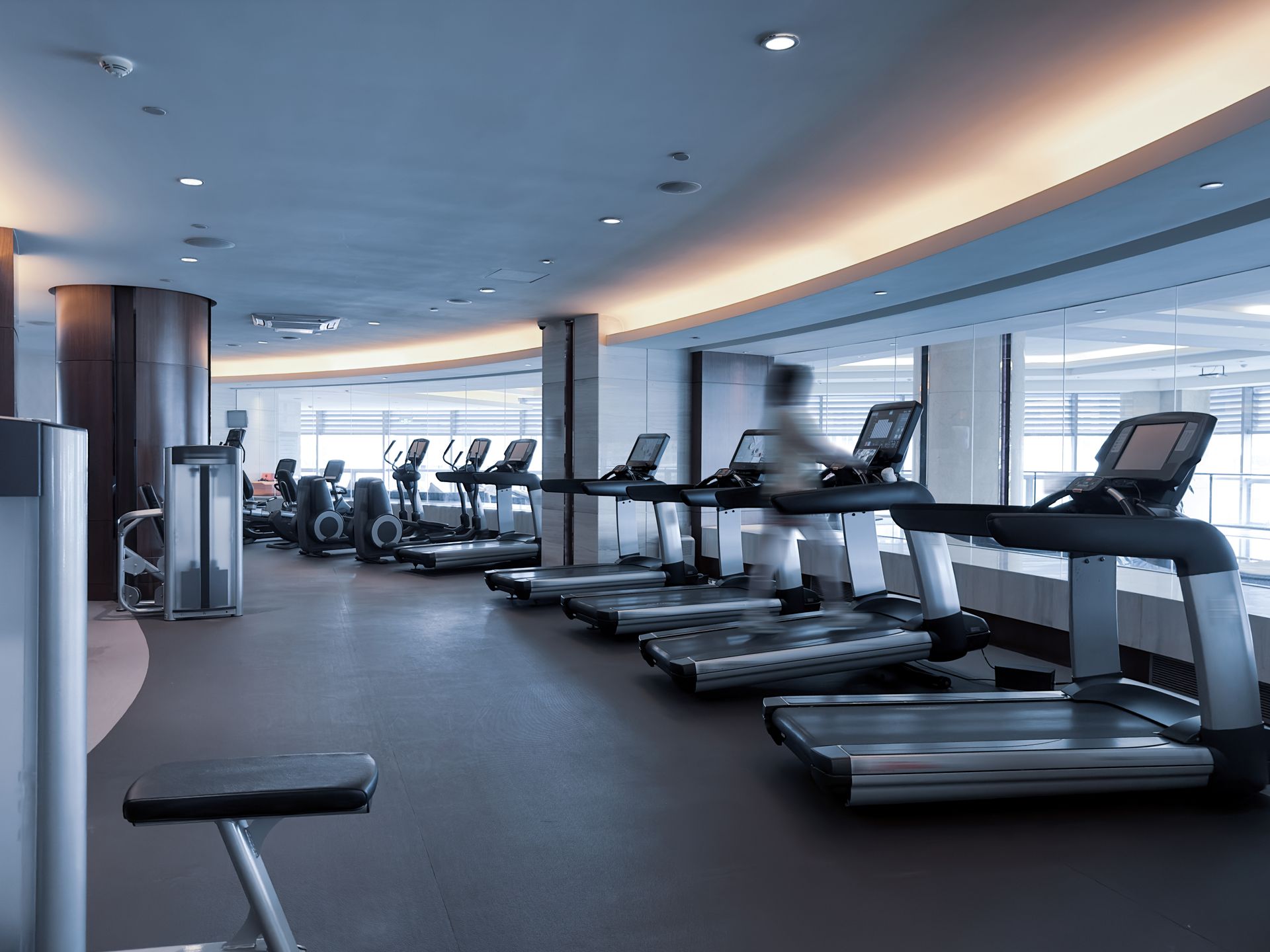A well-lit, modern gym featuring a row of treadmills and fitness equipment on a dark floor in a spacious, neutral room.