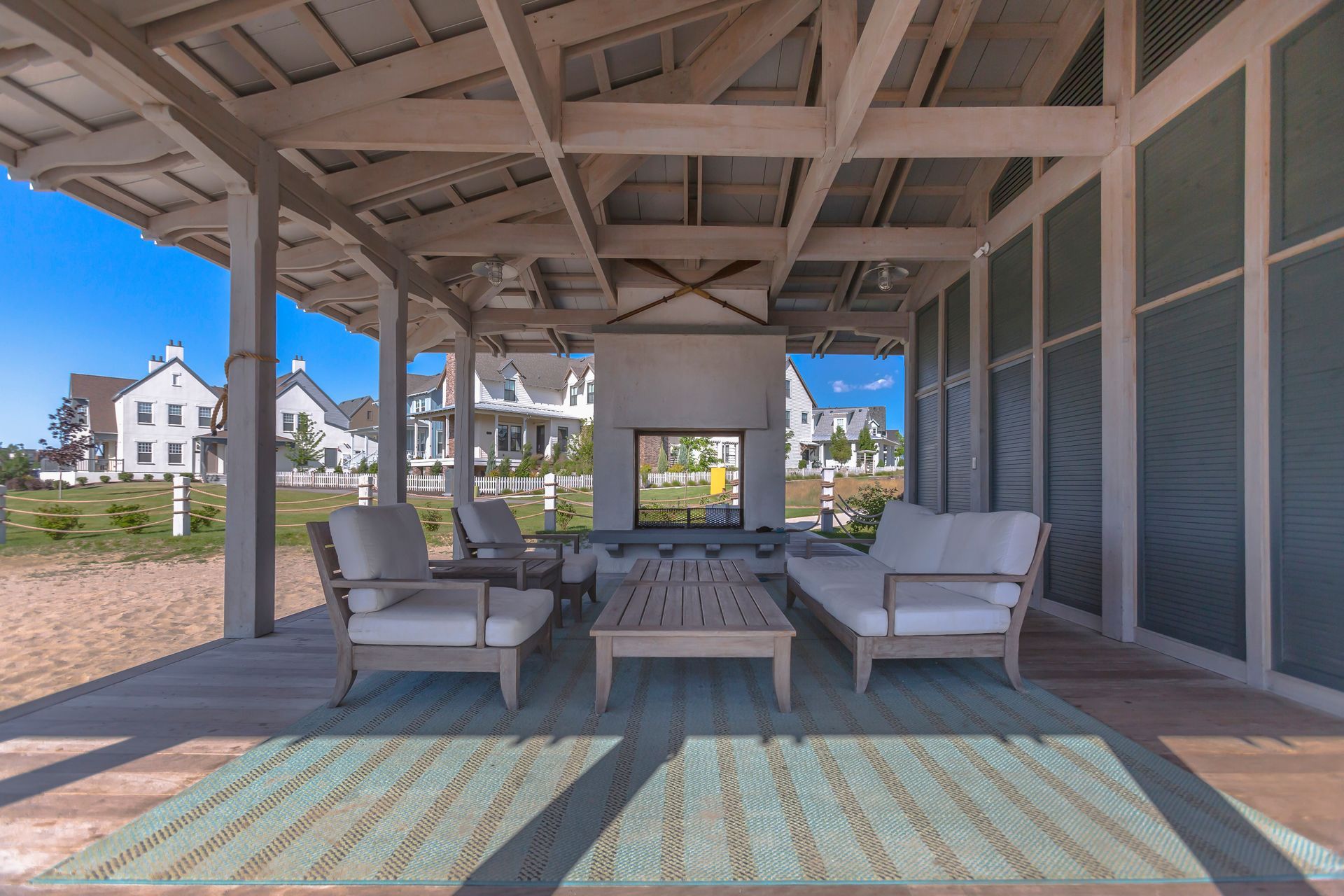 Outdoor lounge area with light-colored furniture under a wooden canopy. A fireplace and houses are visible in the background.