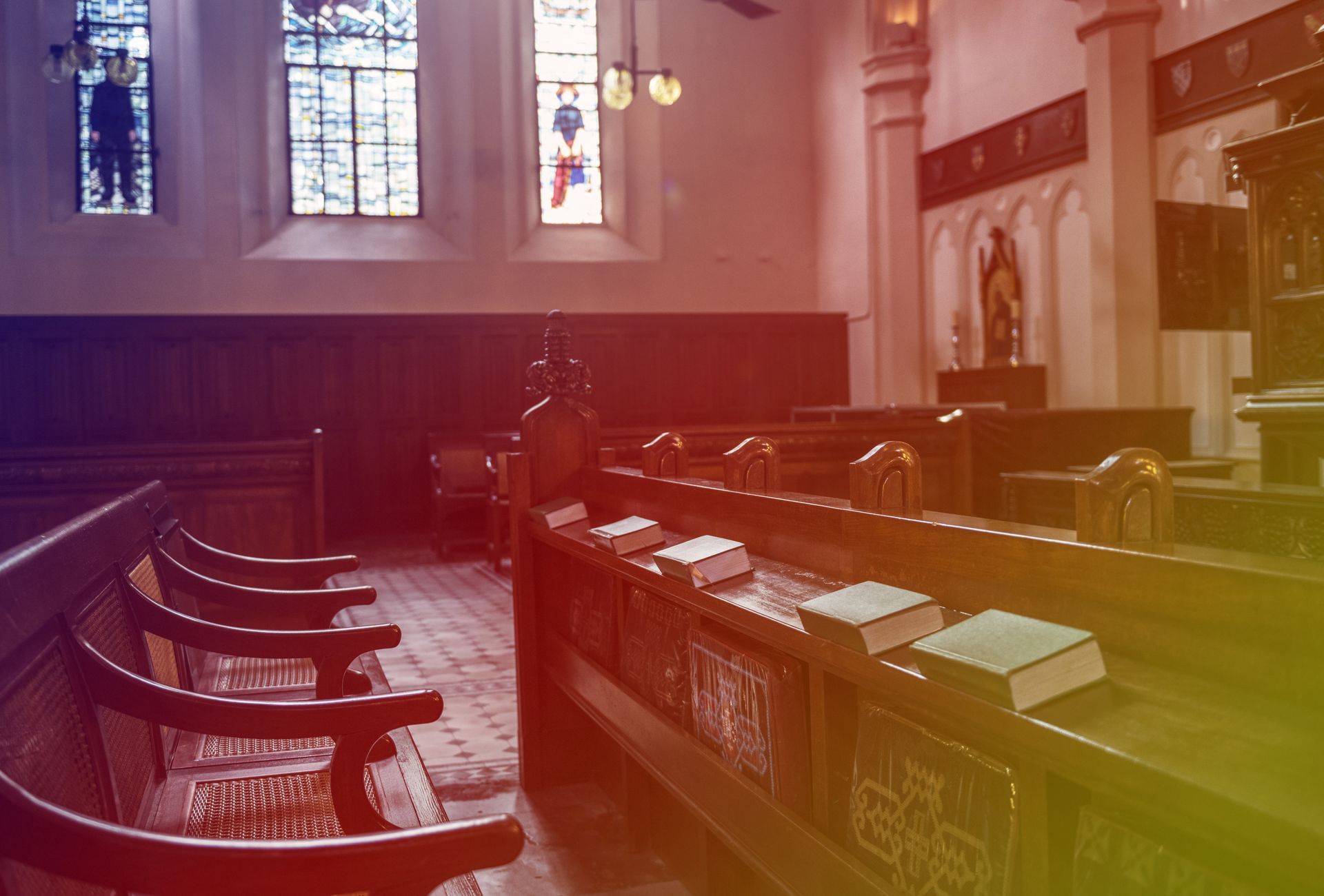 Interior of a church with wooden pews and stained-glass windows. Books rest on the pews.
