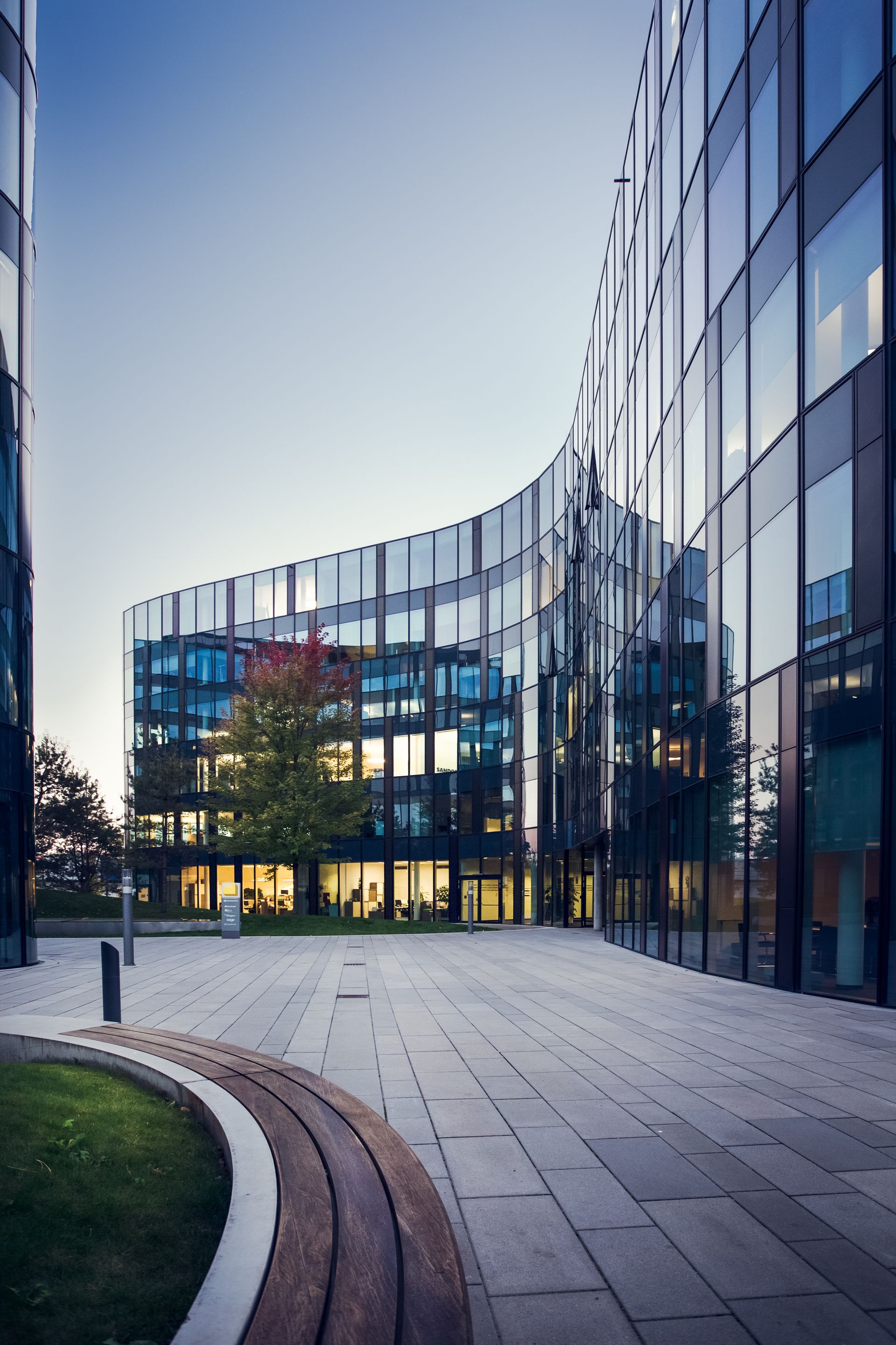 A curved glass office building overlooks a paved plaza with a decorative, arched seating area and a single tree at sunset.
