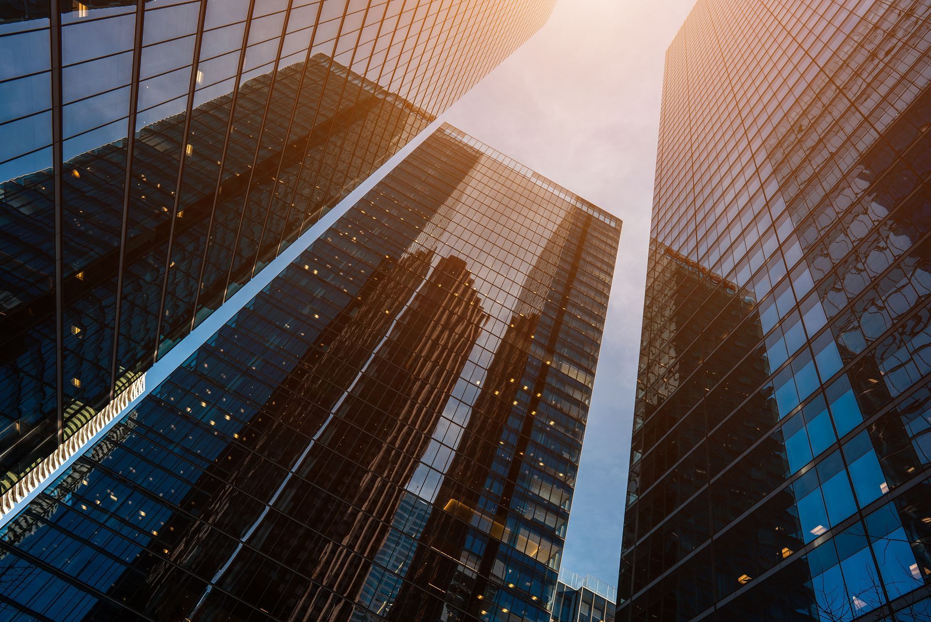 Low-angle view of modern glass skyscrapers reaching toward a bright, sunlit sky.