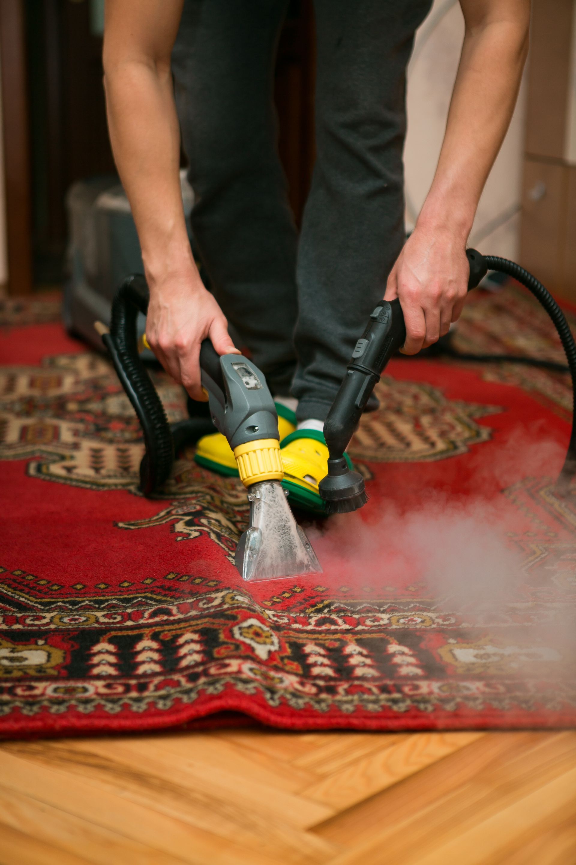 Hands using a steam cleaner on a red patterned rug, with steam rising over a wooden floor edge