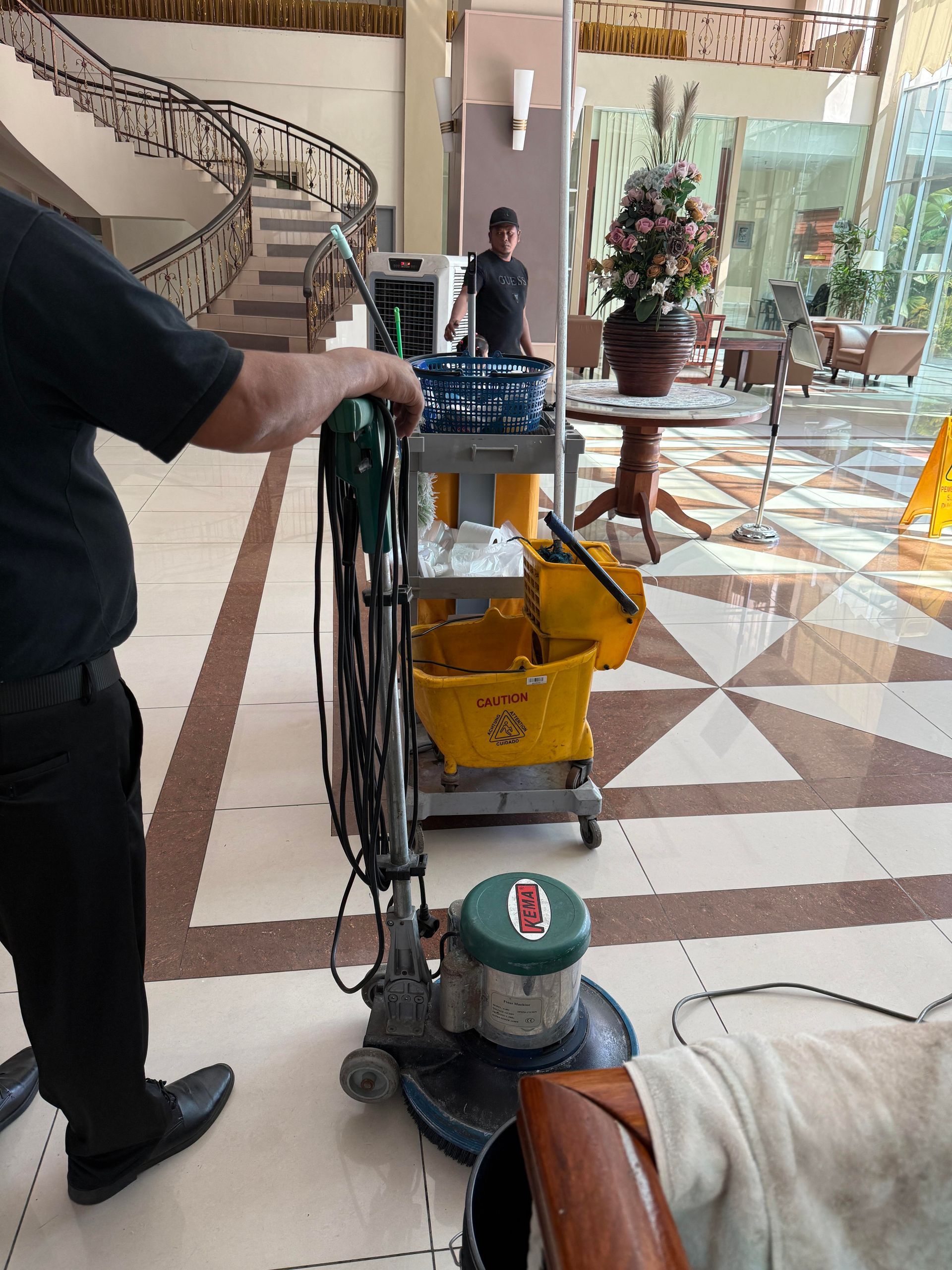 Janitor cleaning a hotel lobby floor with a floor machine and yellow bucket near a staircase