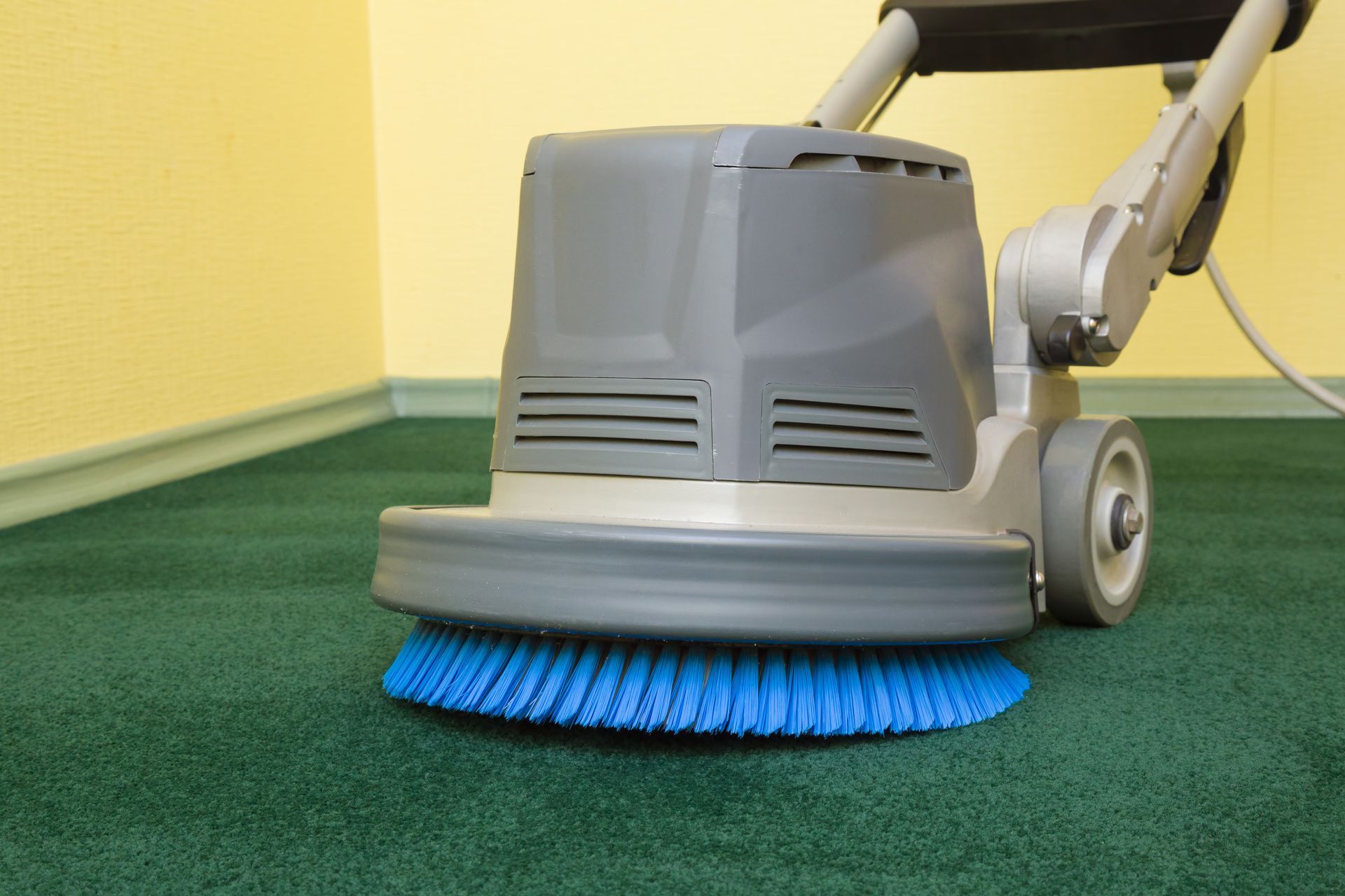 A floor buffer with a blue scrubbing brush cleaning a green carpet in a corner of a room.
