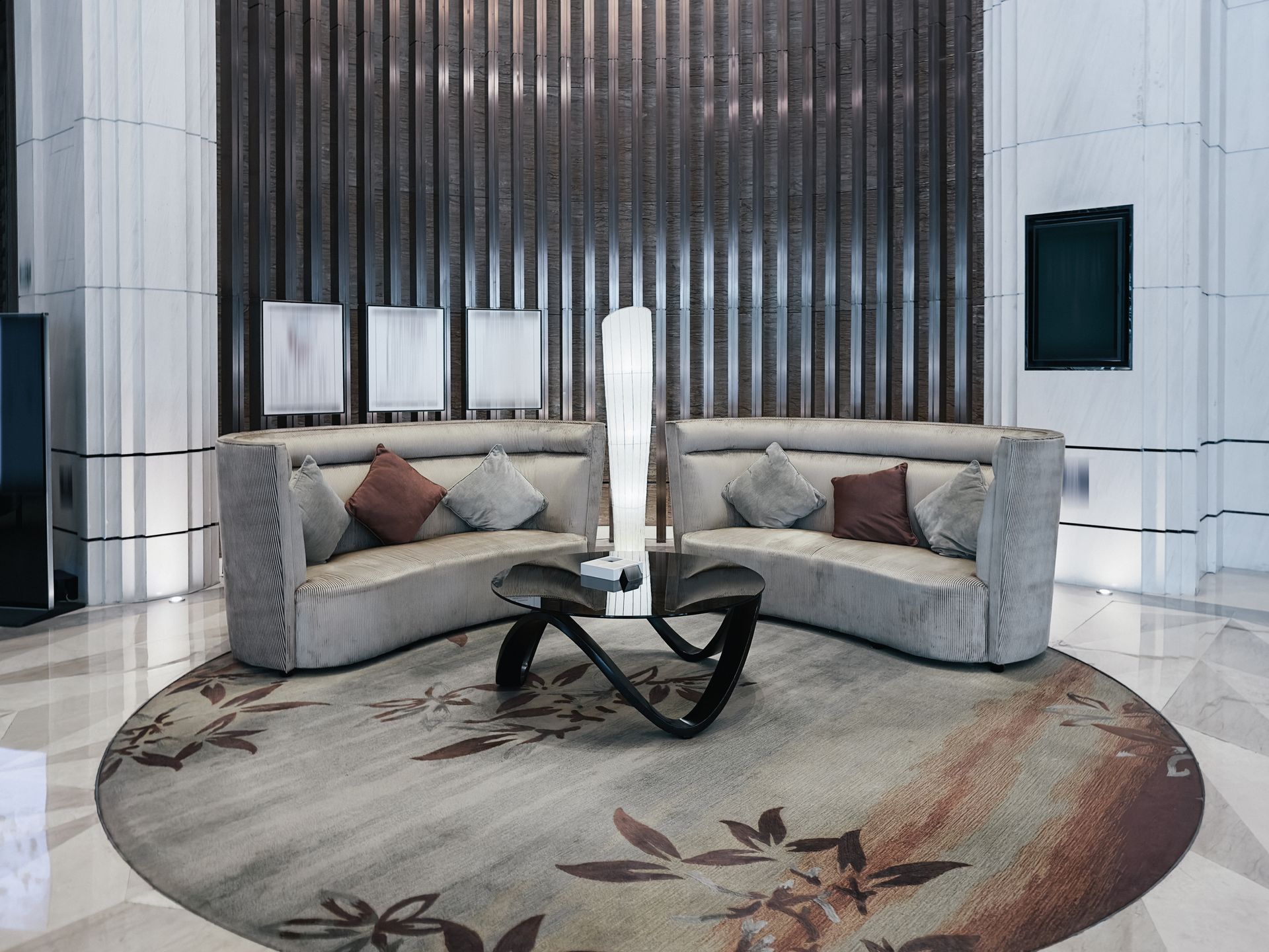 Two curved grey sofas face each other with a modern coffee table on a circular patterned rug in a minimalist lobby.