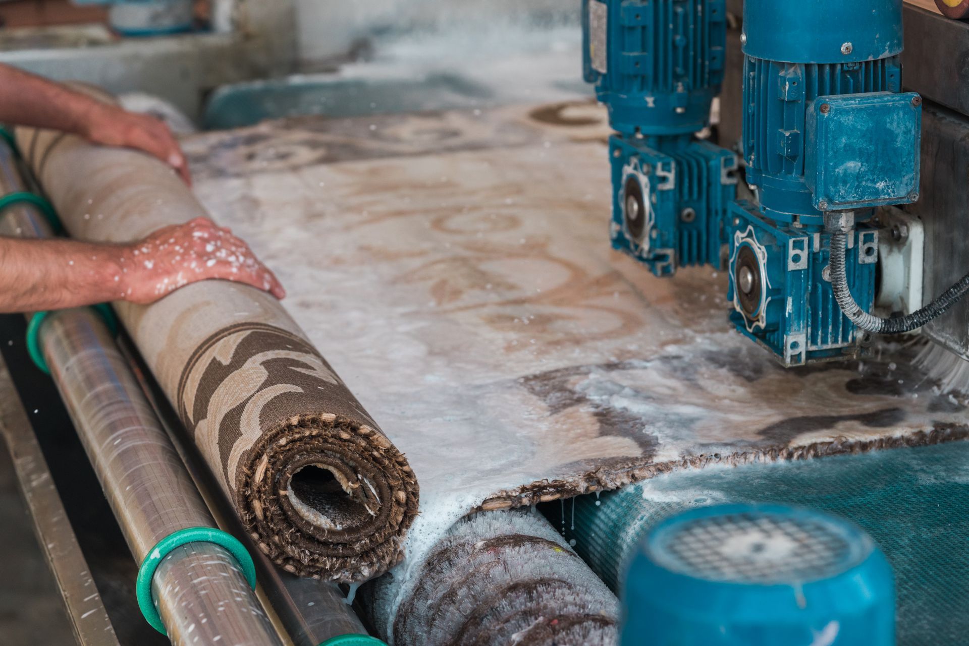 A person cleans a rolled patterned rug using a motorized industrial washing machine with soapy water.