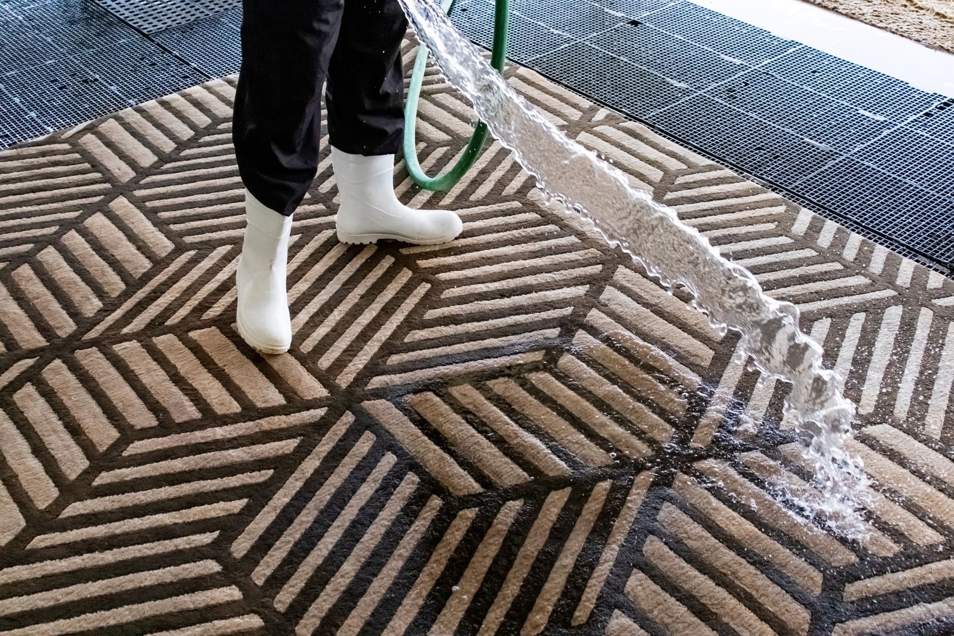 A person in white boots uses a hose to spray water onto a geometric-patterned brown and cream carpet on a mesh surface.