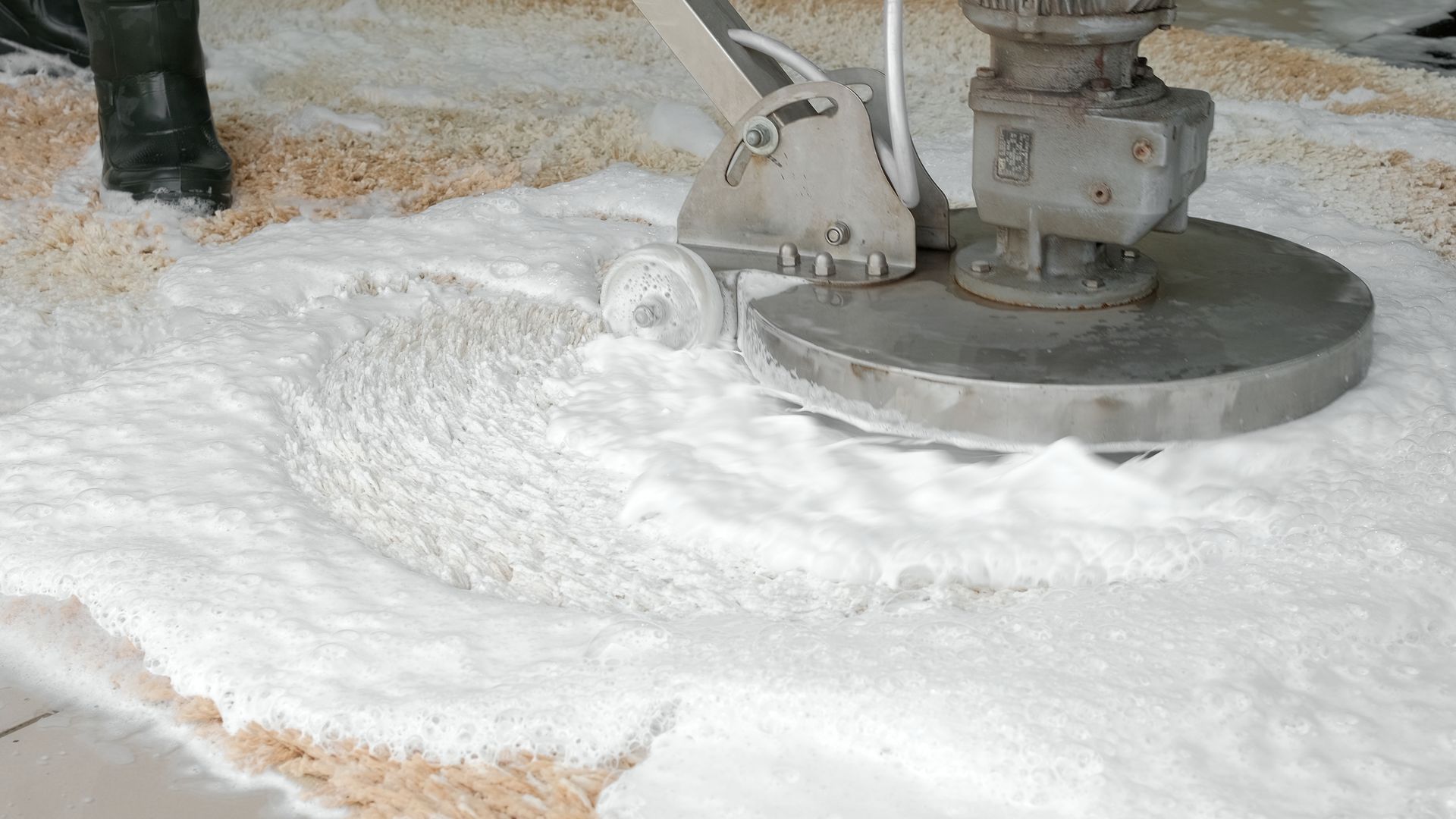 A commercial floor scrubber machine spreading white foam over a carpeted floor.