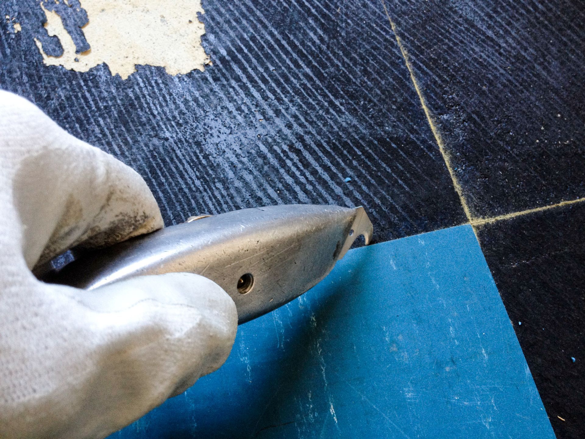 A gloved hand holds a hook-bladed utility knife, scoring a seam between black and blue floor tiles.