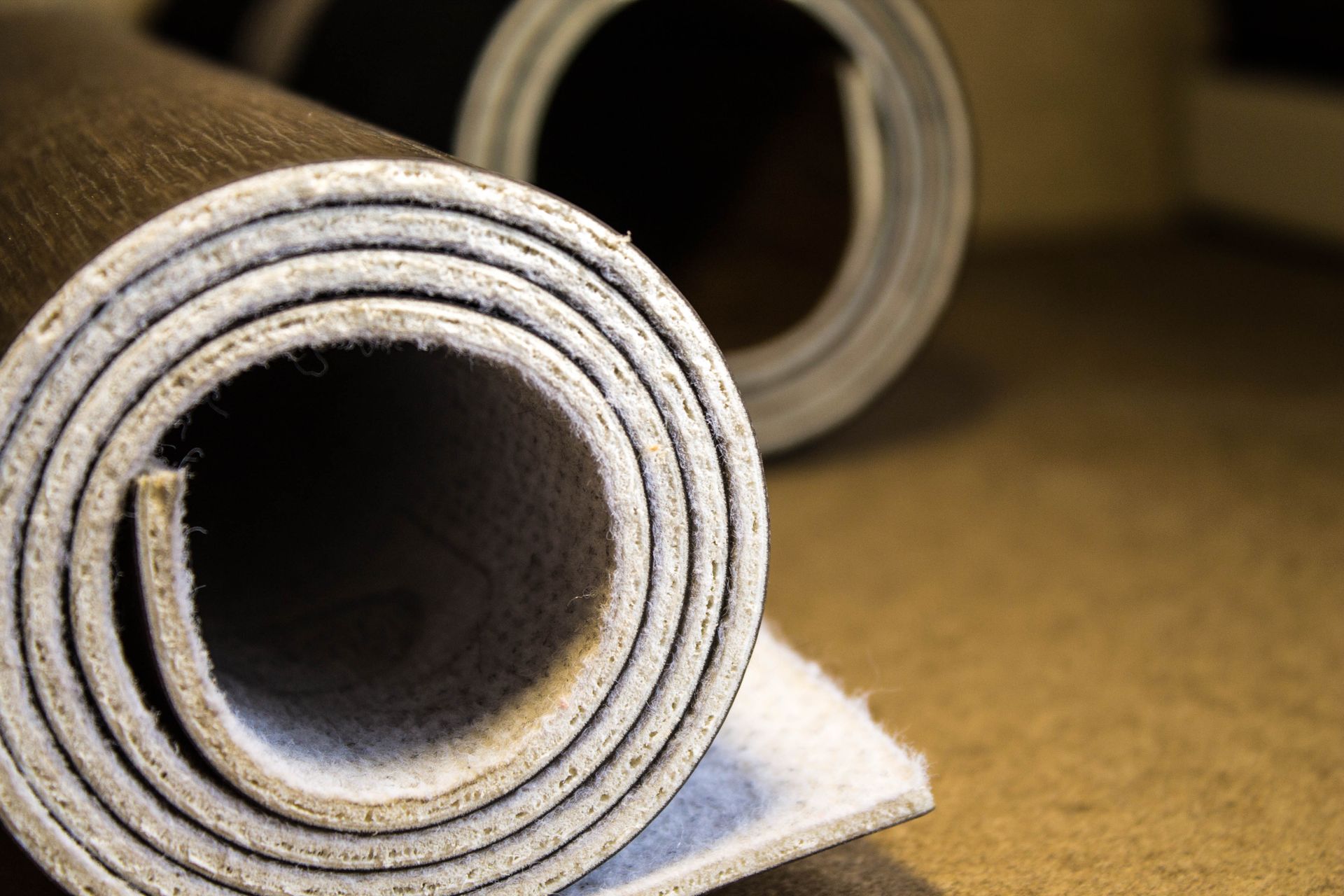 Two rolls of wood-patterned vinyl flooring with thick, light-colored felt backing, resting on a flat, neutral surface.