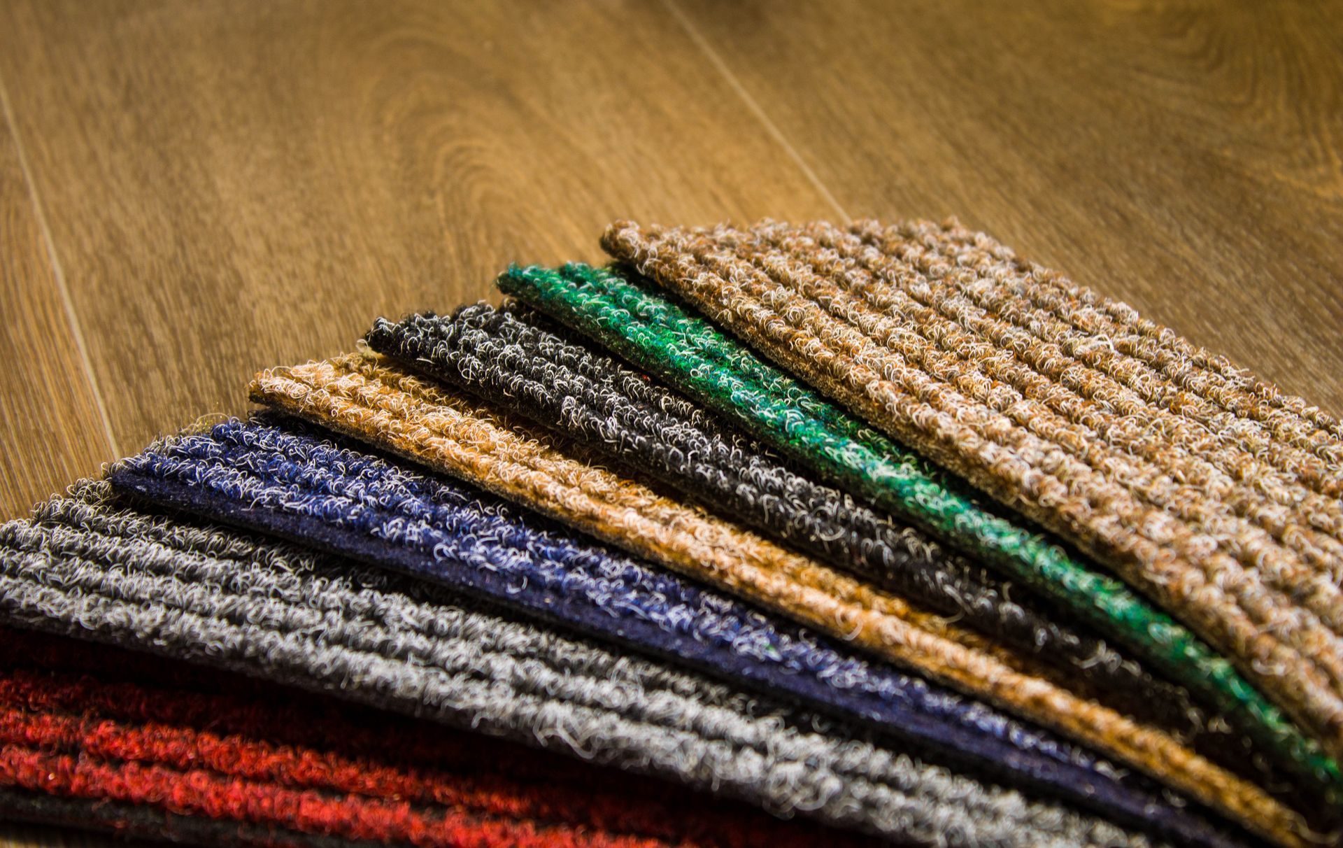 A fan of carpet samples in various colors—red, grey, blue, tan, charcoal, green, and beige—laid on a wooden floor.