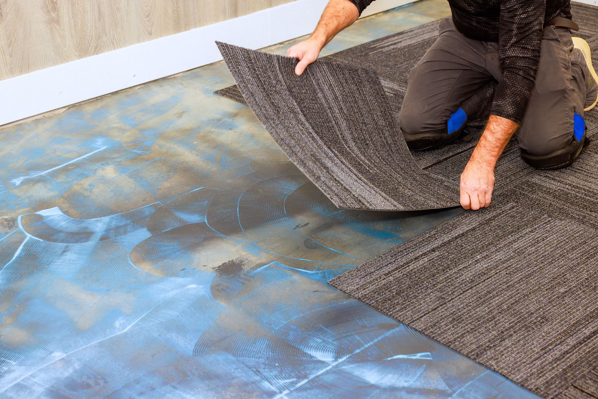 A person kneels on a floor, installing grey carpet tiles onto a subfloor coated with blue adhesive.
