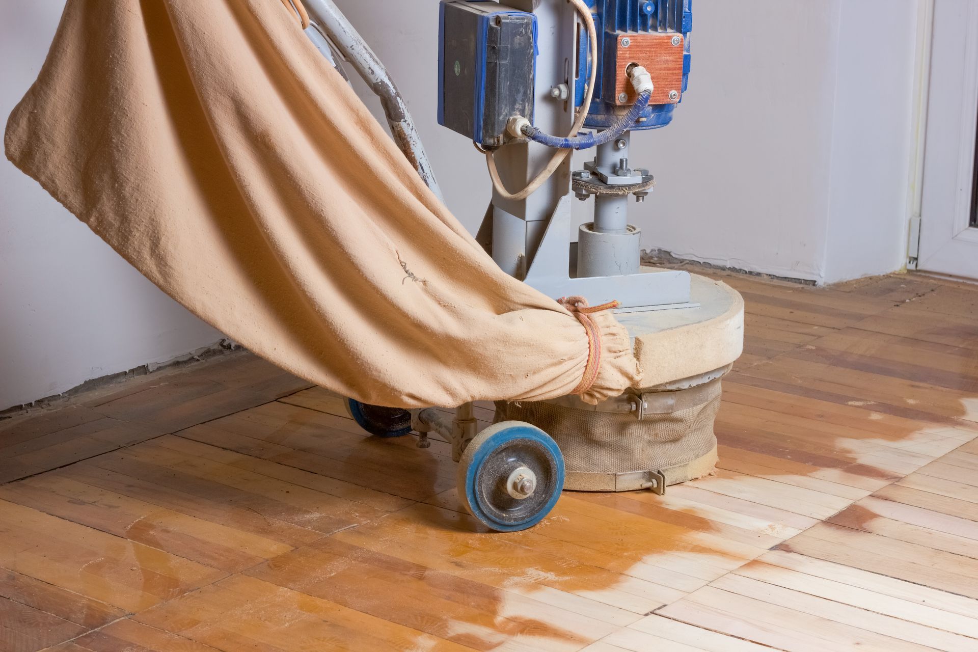 A heavy-duty floor sanding machine with a large dust collection bag operates on a wooden floor inside a room.