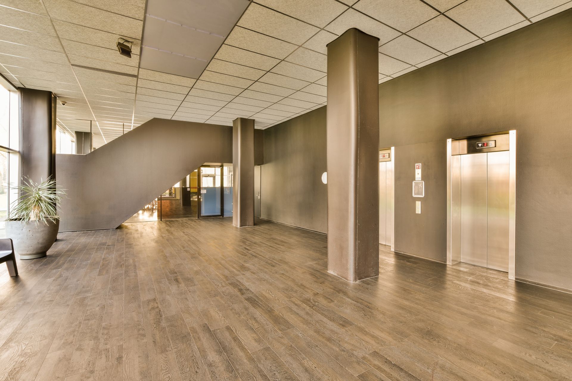 A modern, minimalist building lobby with dark walls, wood-grain flooring, two metal elevators, and a wide staircase.