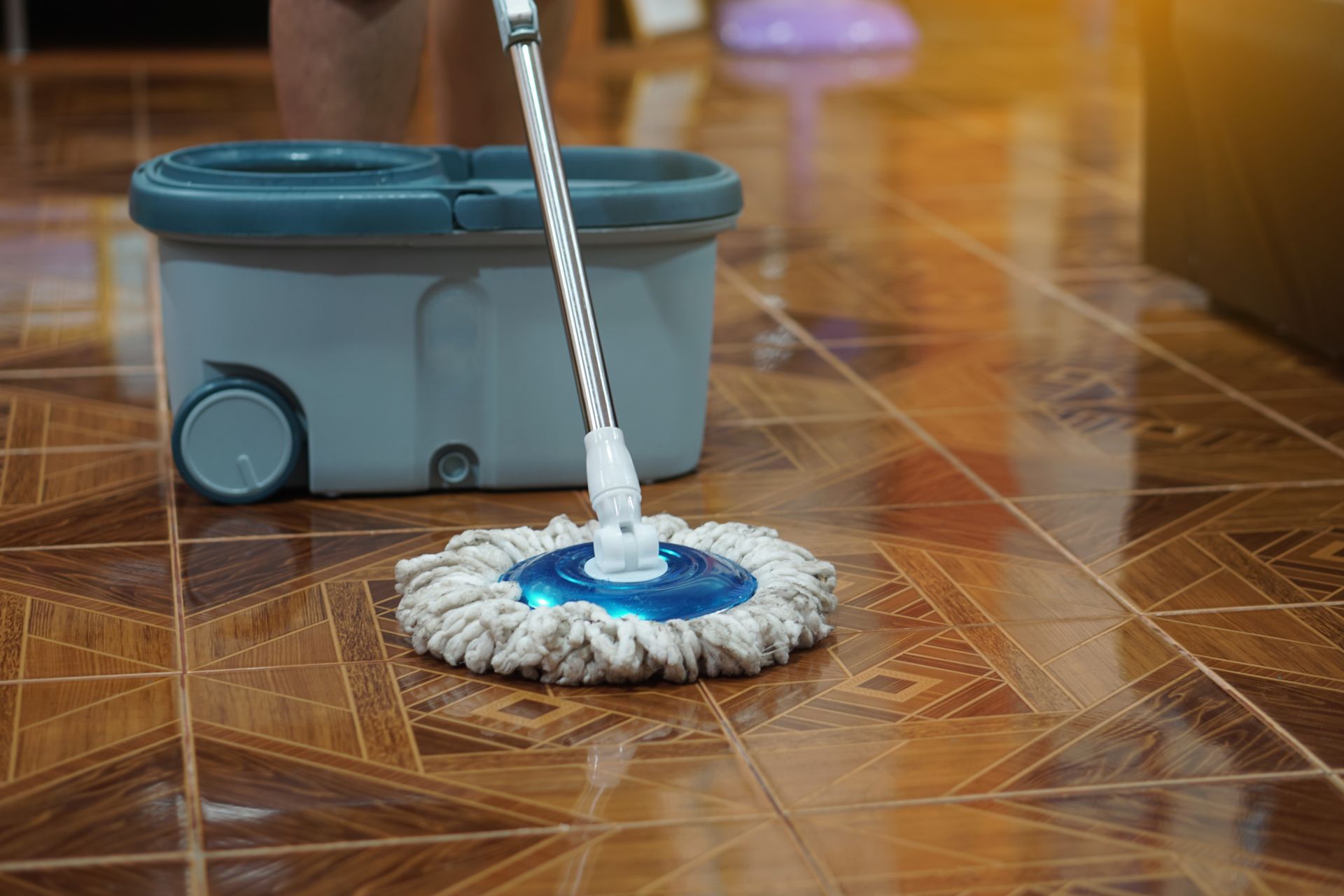 A mop with a circular white head rests on a brown patterned tile floor next to a gray plastic bucket.