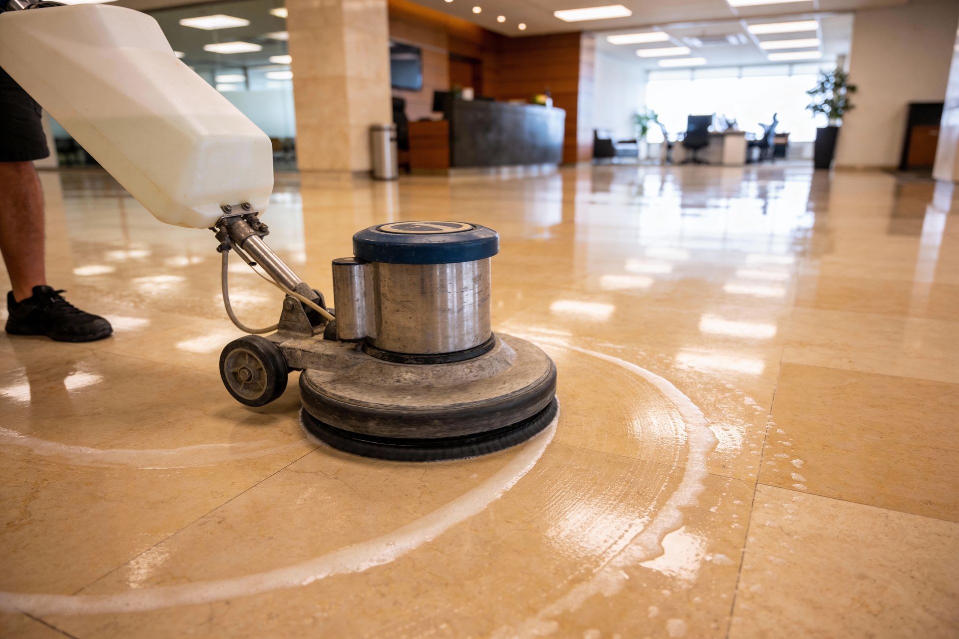 A person uses a floor buffer to polish a shiny, light-colored tile floor in a modern office lobby.