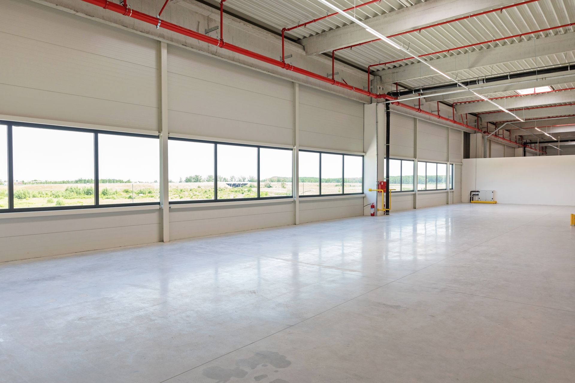 A wide, empty, modern warehouse space with light gray concrete floors, white walls, and large windows looking outside.