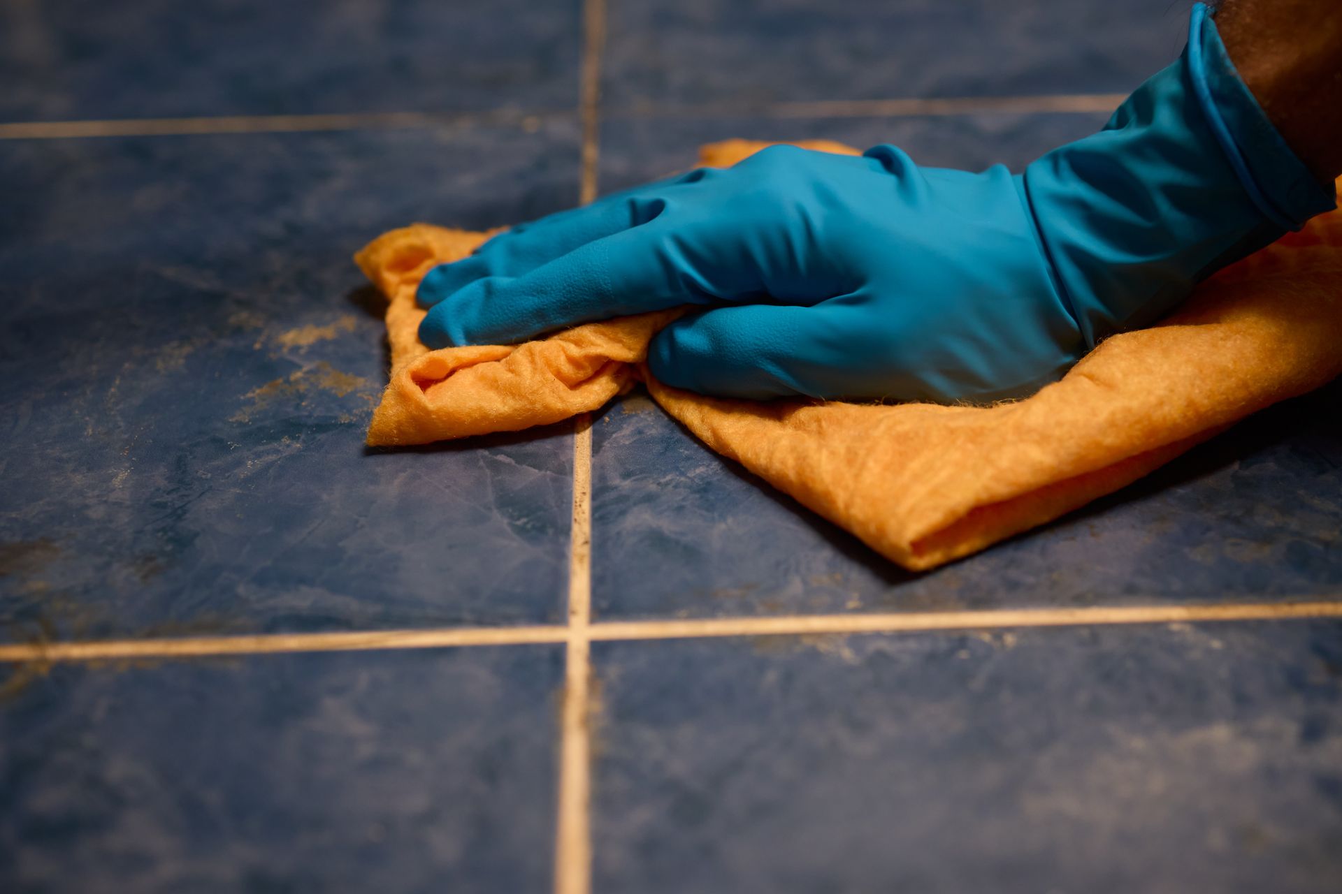 A hand in a blue rubber glove uses an orange cloth to wipe a dark blue tiled floor.