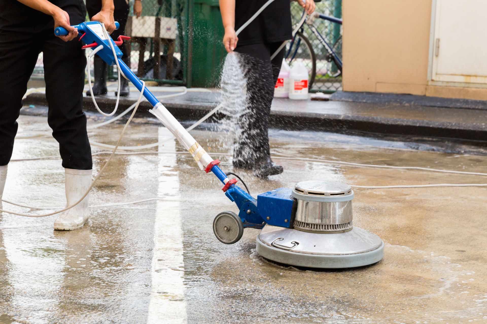 Two people using a floor cleaning machine, spraying water, on a concrete surface.
