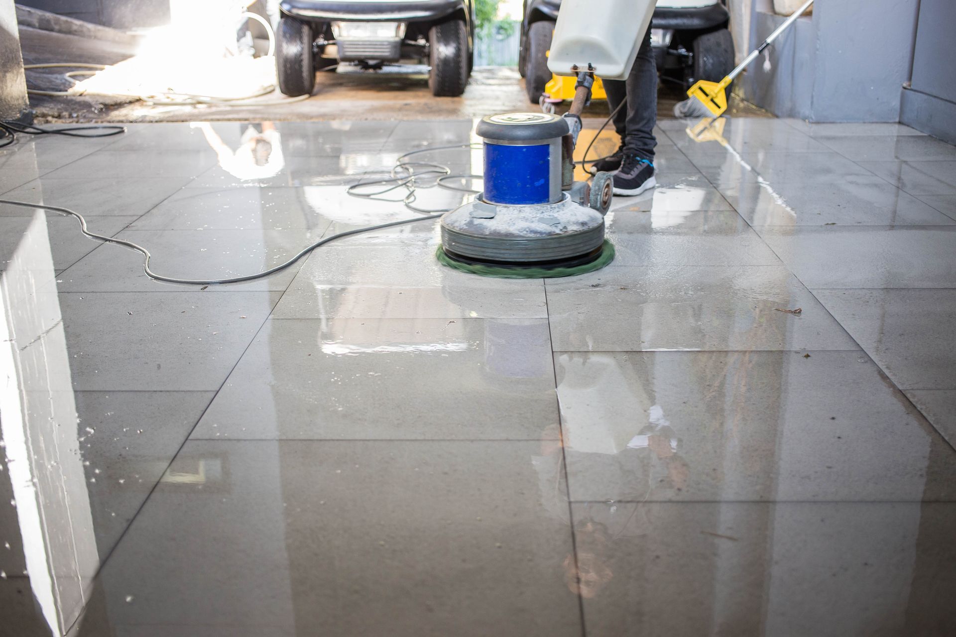 A person uses a floor buffer machine to polish shiny, wet gray tiles in a garage with parked vehicles.