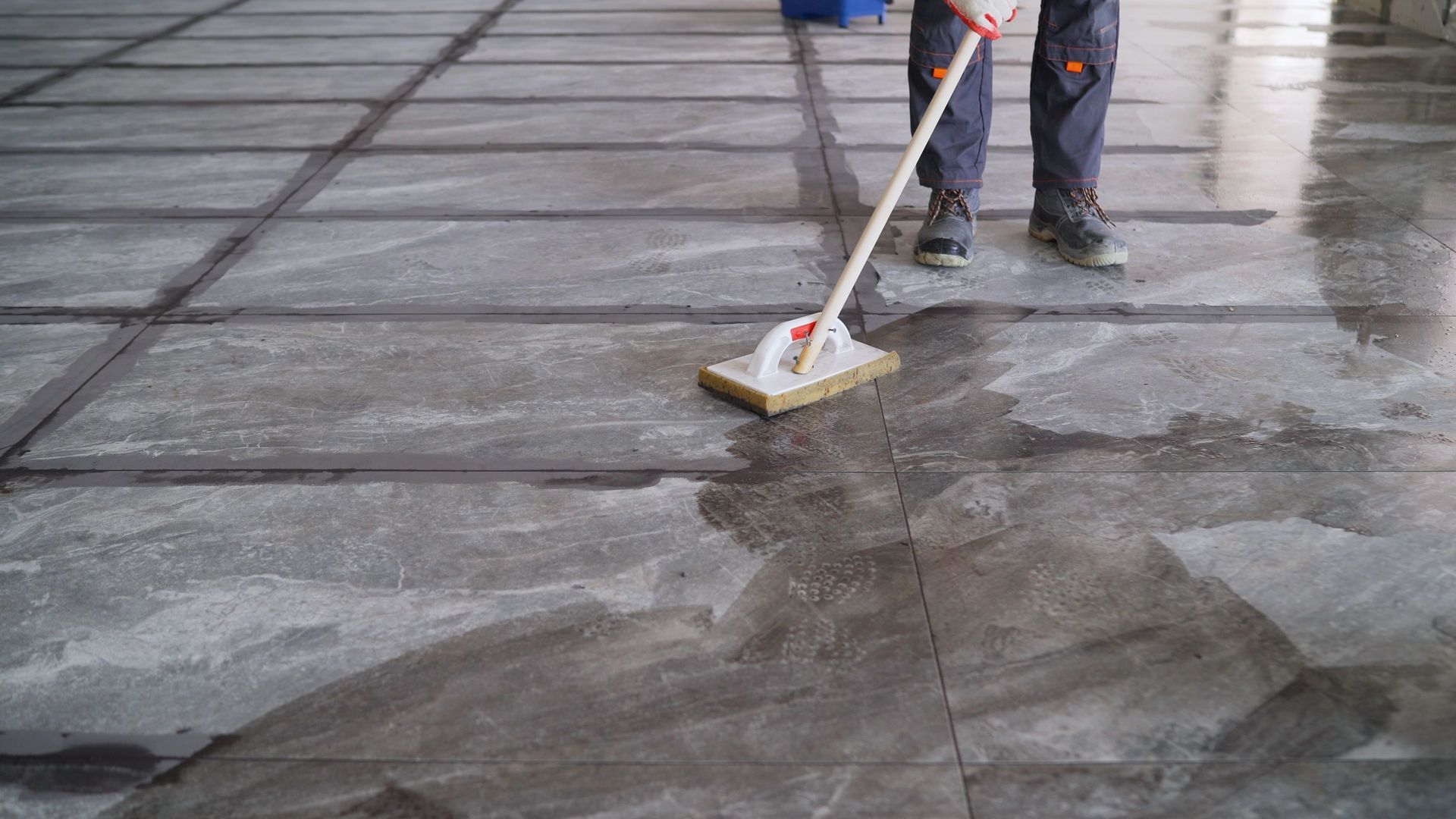 A worker uses a long-handled grout float to smooth mortar over newly laid dark stone tiles on a large floor.