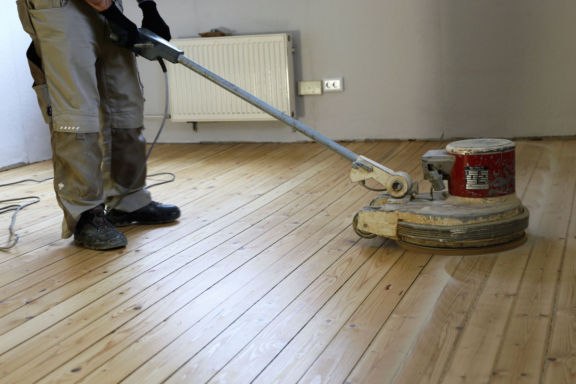 A person uses a floor sanding machine on a wooden floor in a room.