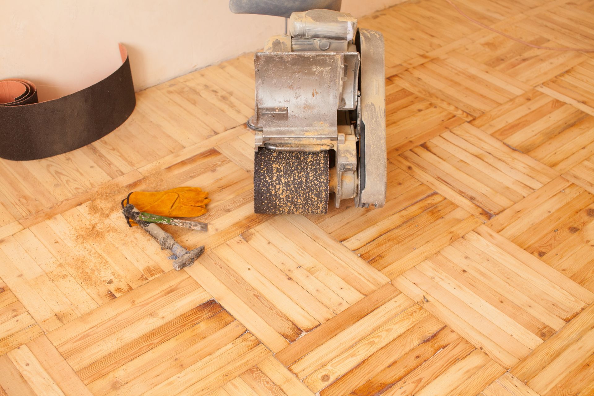 A heavy-duty floor sander sits on unfinished parquet wood flooring near a pair of work gloves and a metal tool.