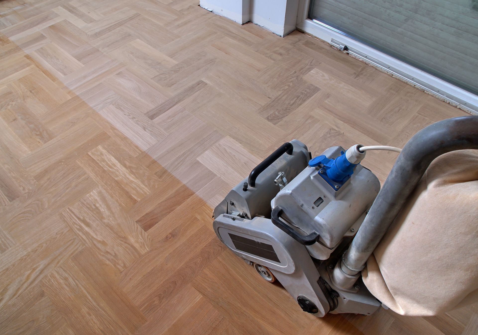 A floor sander machine sits on a partially sanded herringbone wood floor, showing the contrast between old and new wood.