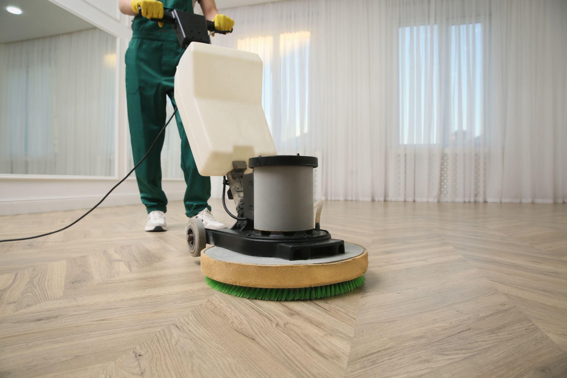 A person in a green uniform uses a floor buffer machine on a light-colored wooden floor in a brightly lit room.
