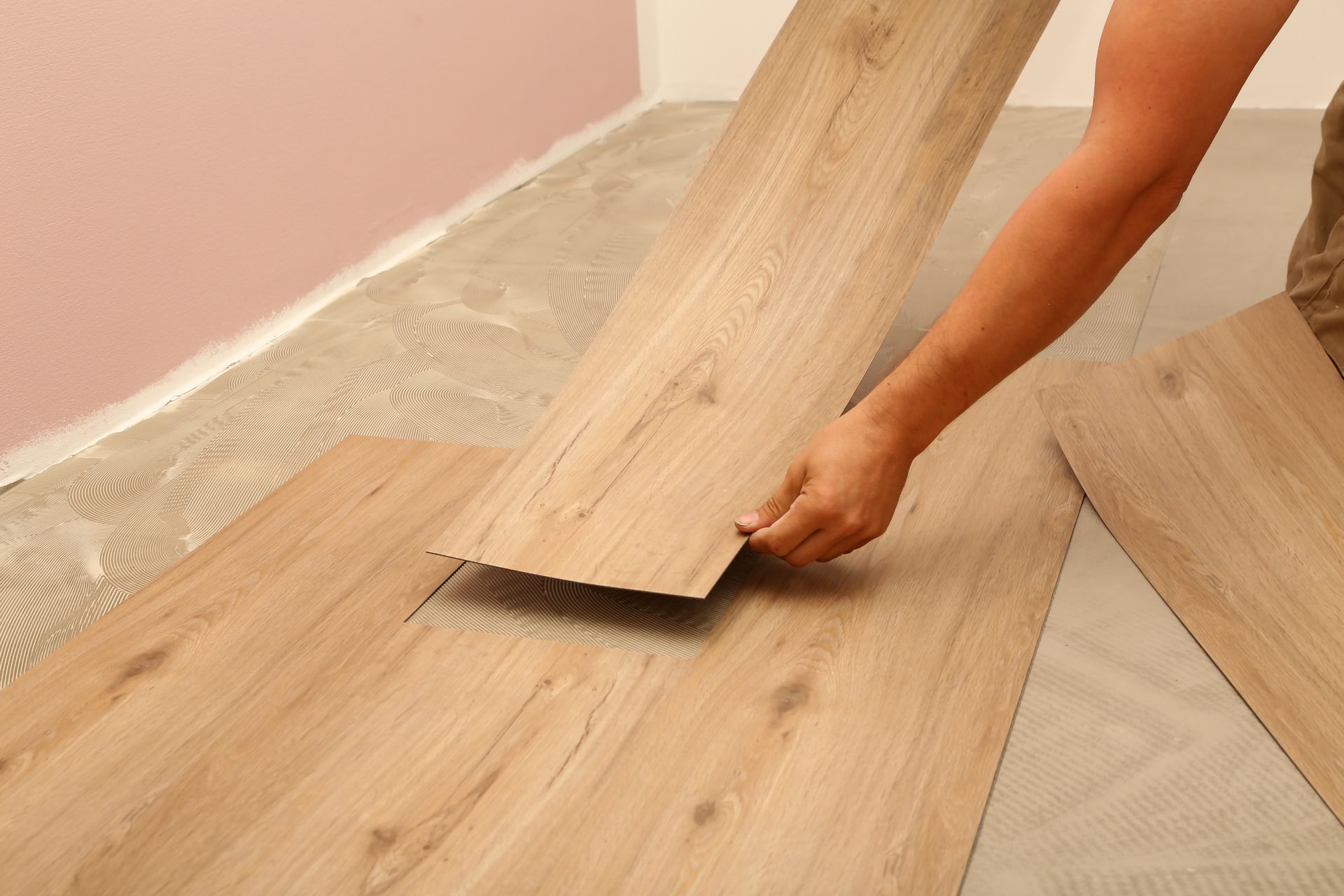 A person installing wood-look vinyl plank flooring over an underlayment near a light pink wall.