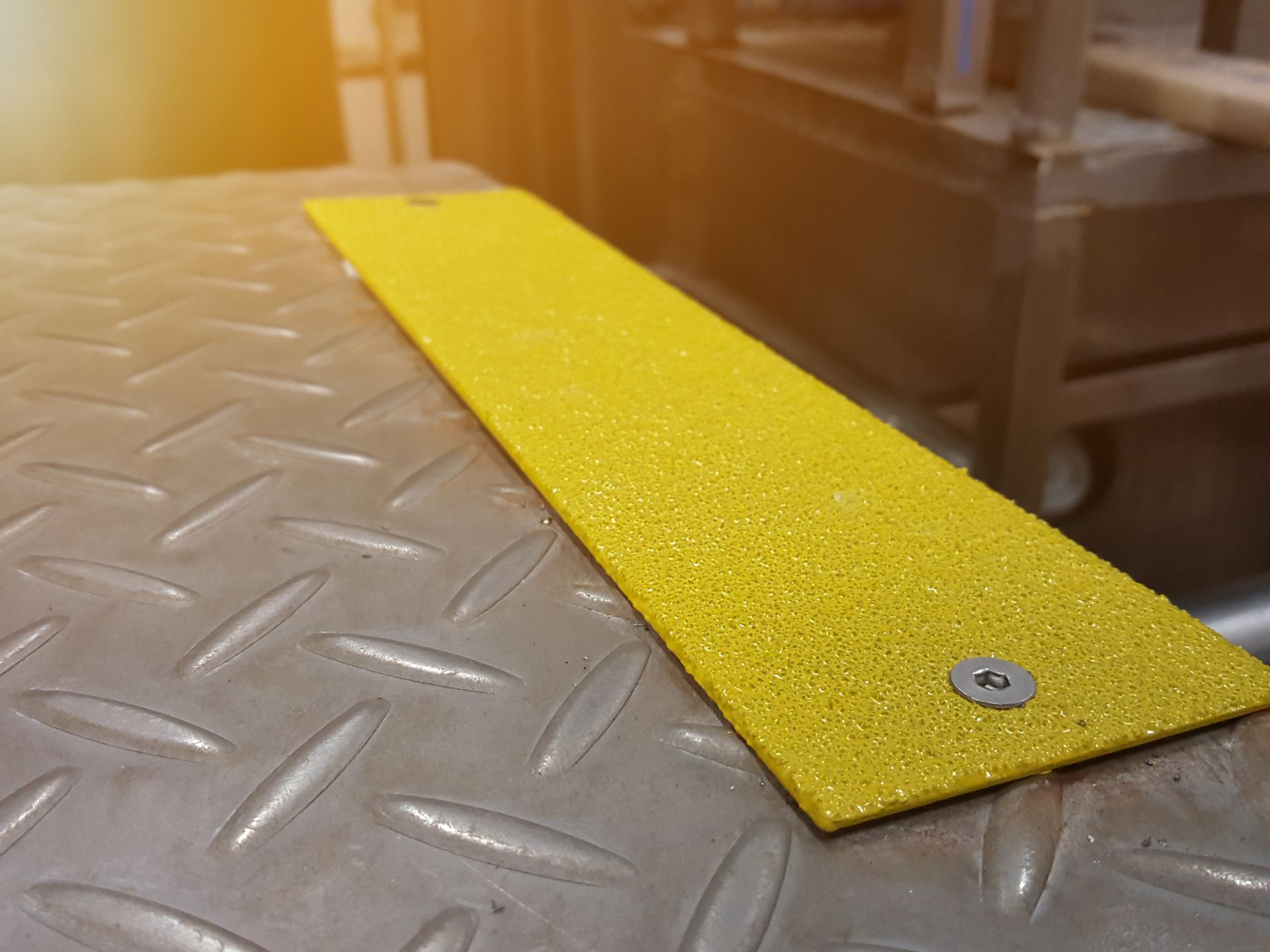 A bright yellow, textured anti-slip safety strip mounted with silver screws onto a gray metal diamond plate surface.