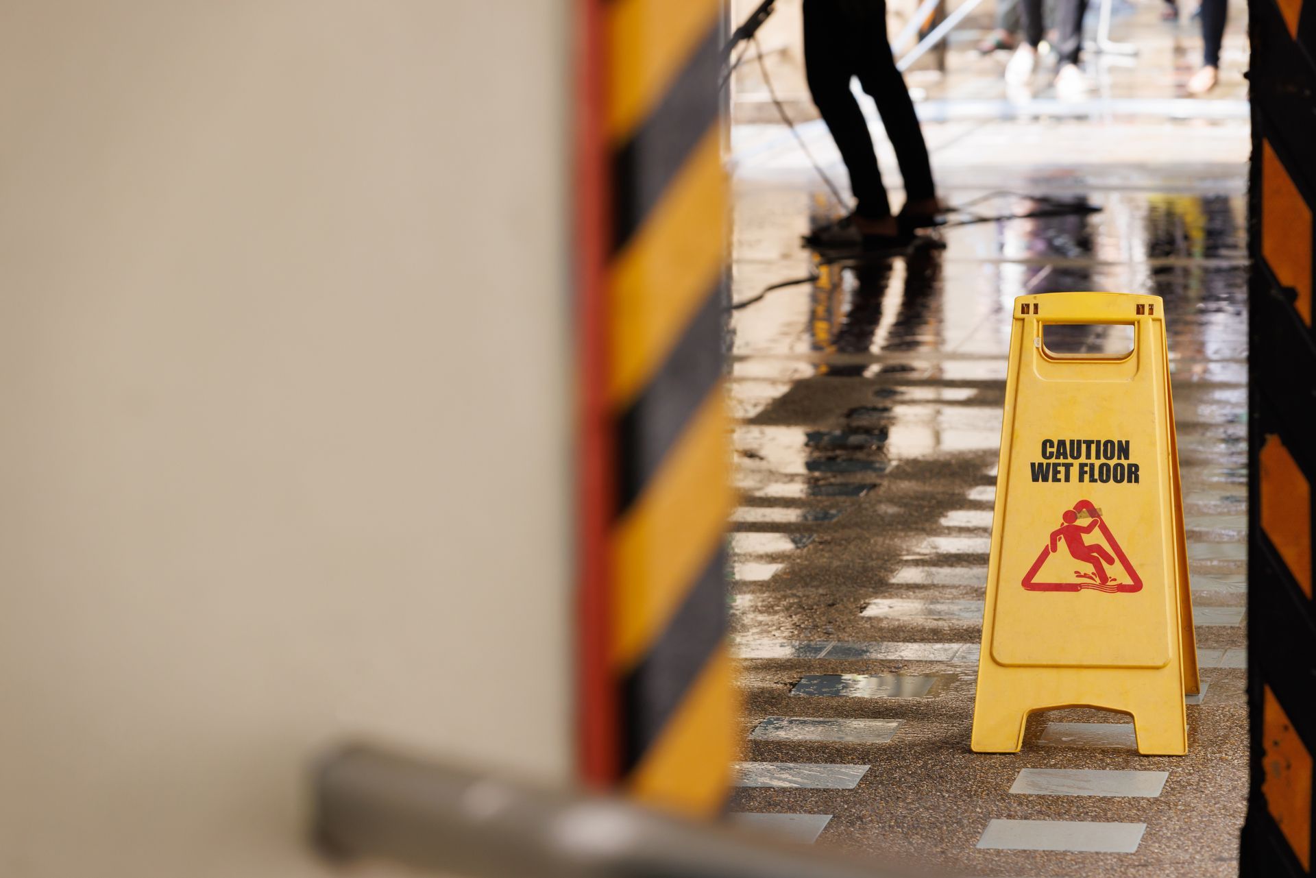 A yellow wet floor caution sign stands on a reflective, damp indoor floor between two black and yellow striped pillars.