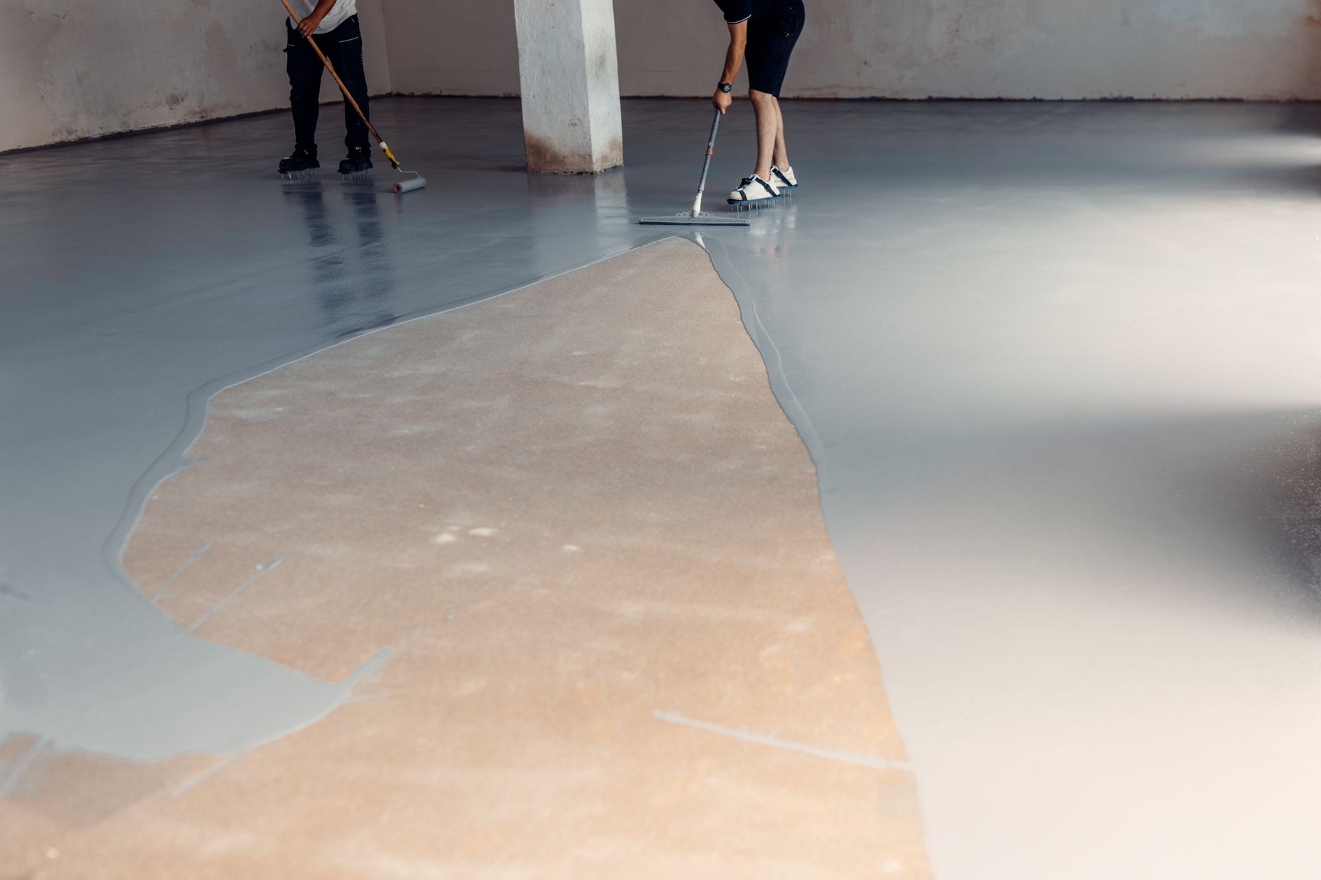 Two workers use long-handled tools to apply smooth, gray epoxy coating to a concrete floor in an unfinished room.
