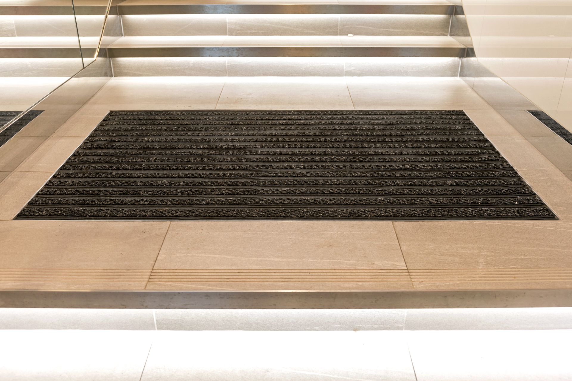 A rectangular dark gray ribbed entryway mat sits on a tiled floor in front of a staircase with lit steps.