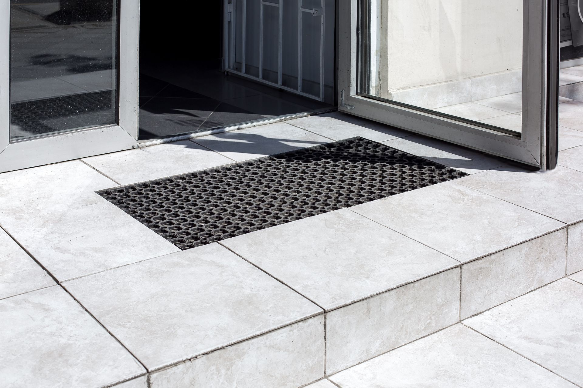 A dark, rectangular outdoor floor mat with a geometric pattern, centered on light-colored tile steps outside a doorway.