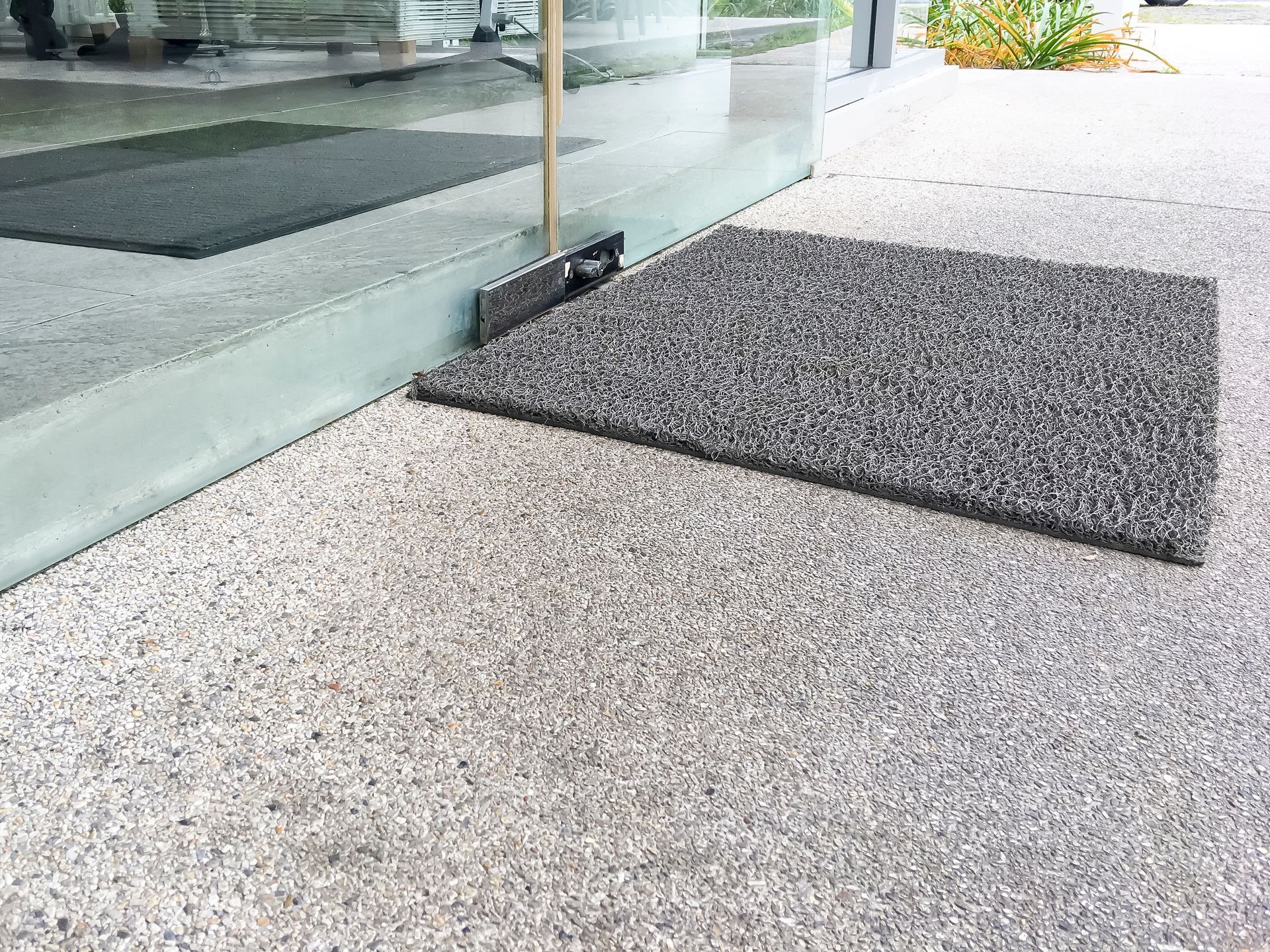 A grey, textured doormat placed on a concrete entrance, positioned next to a glass door.