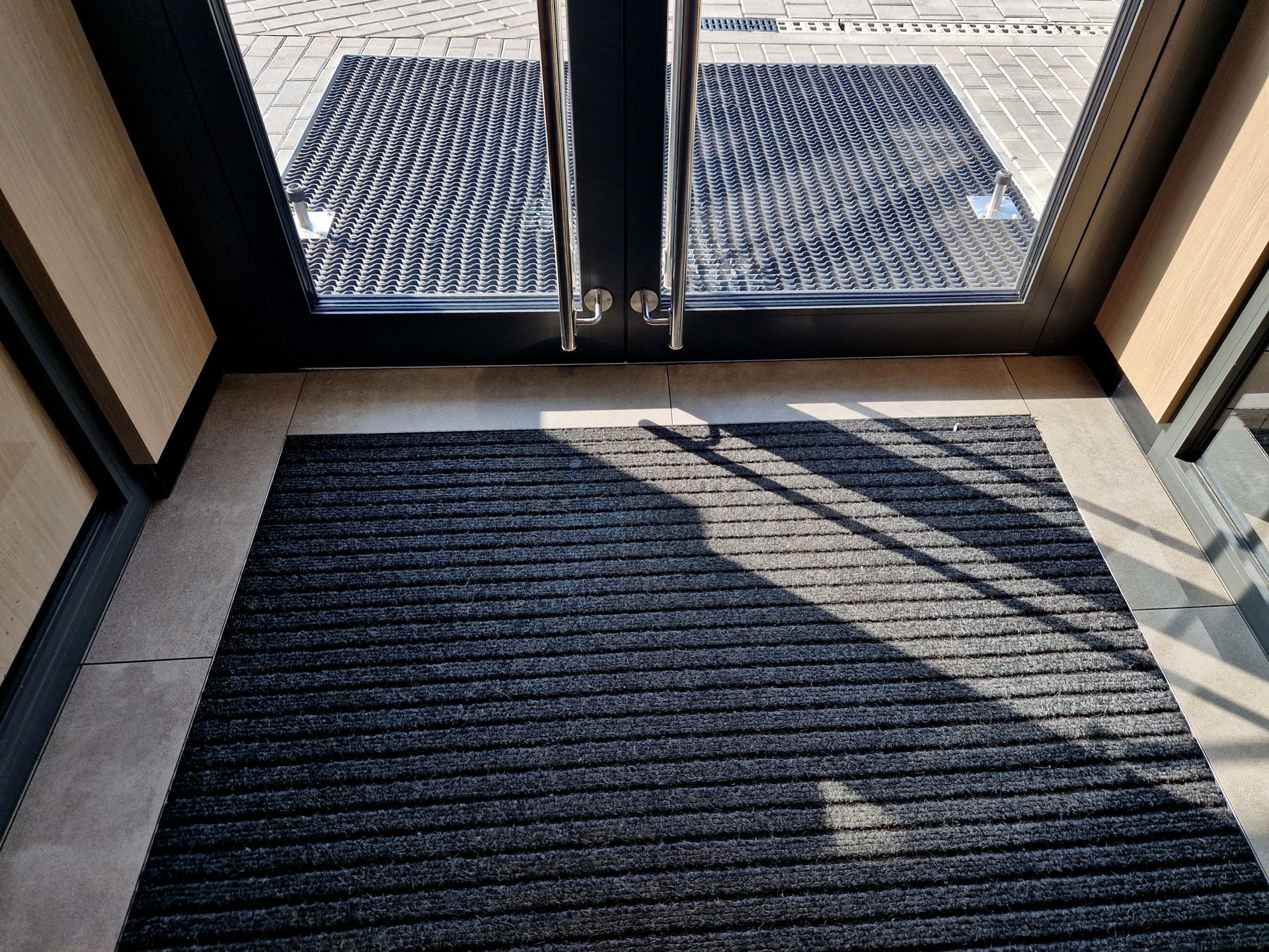 An indoor dark-ribbed mat placed on a tiled floor in front of glass double doors leading to an outdoor walkway.