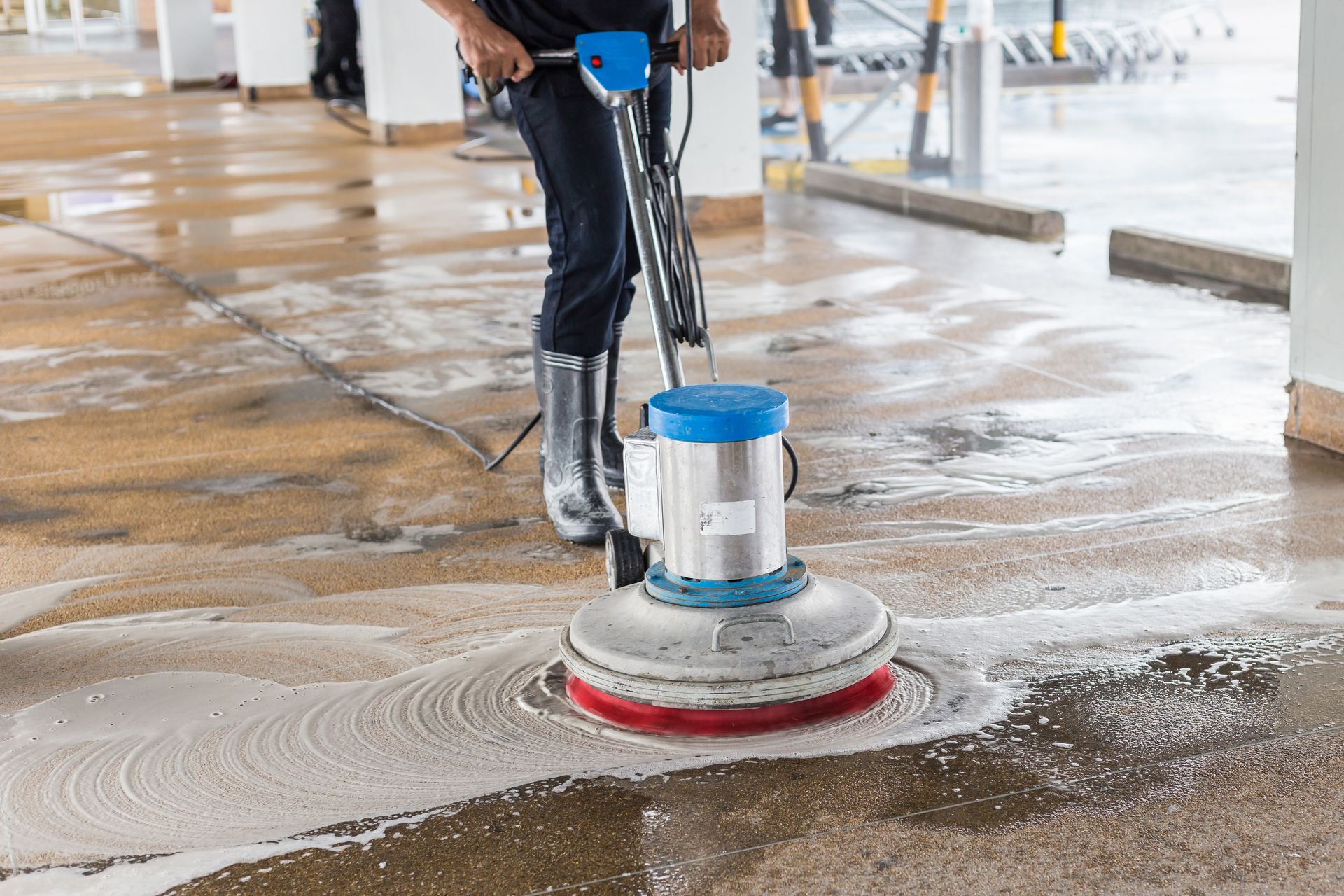 A person in dark clothing and gray rubber boots uses a blue and silver floor buffer to clean a concrete floor.