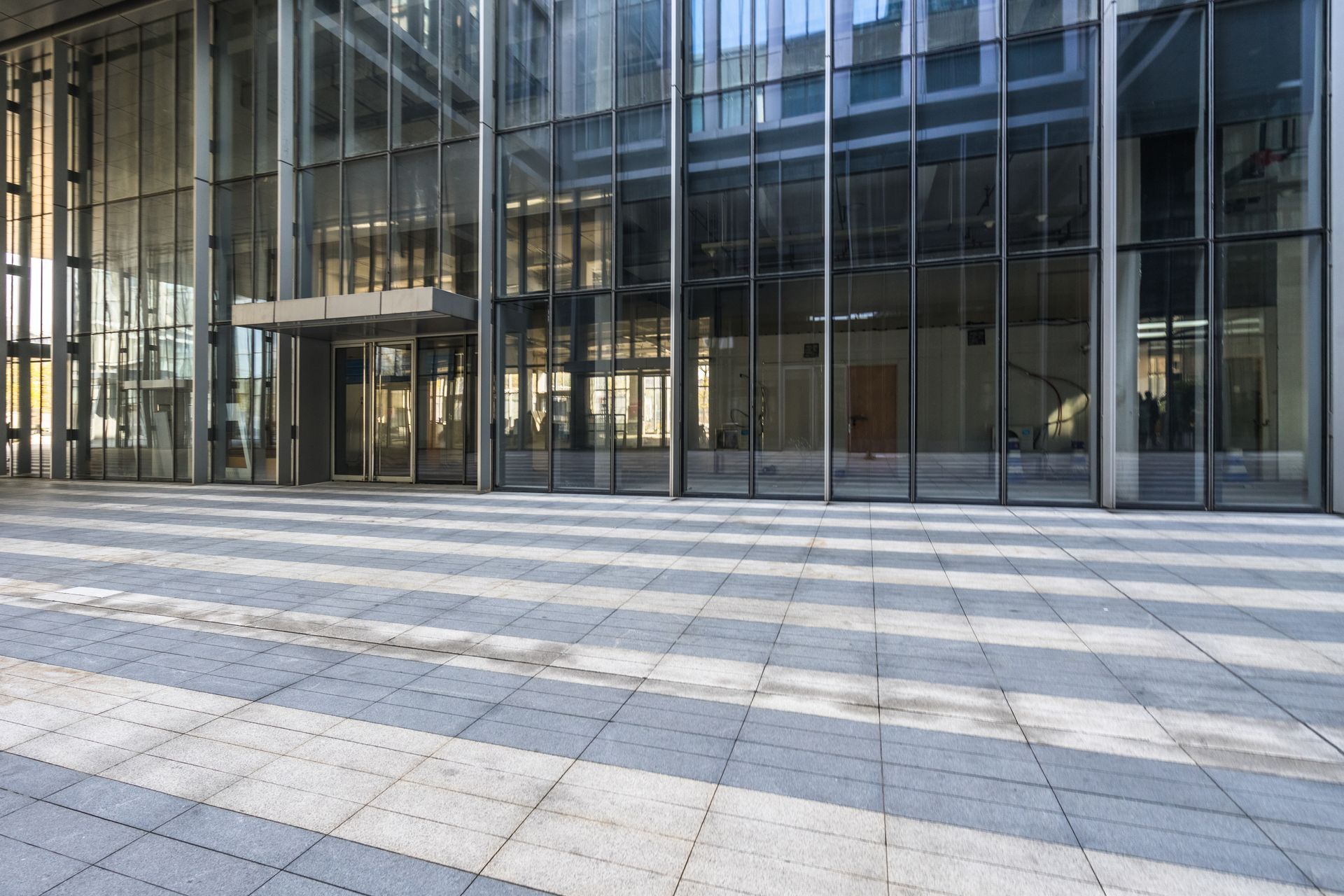 Modern glass facade of an office building exterior with a tiled plaza featuring a pattern of light and dark stripes.