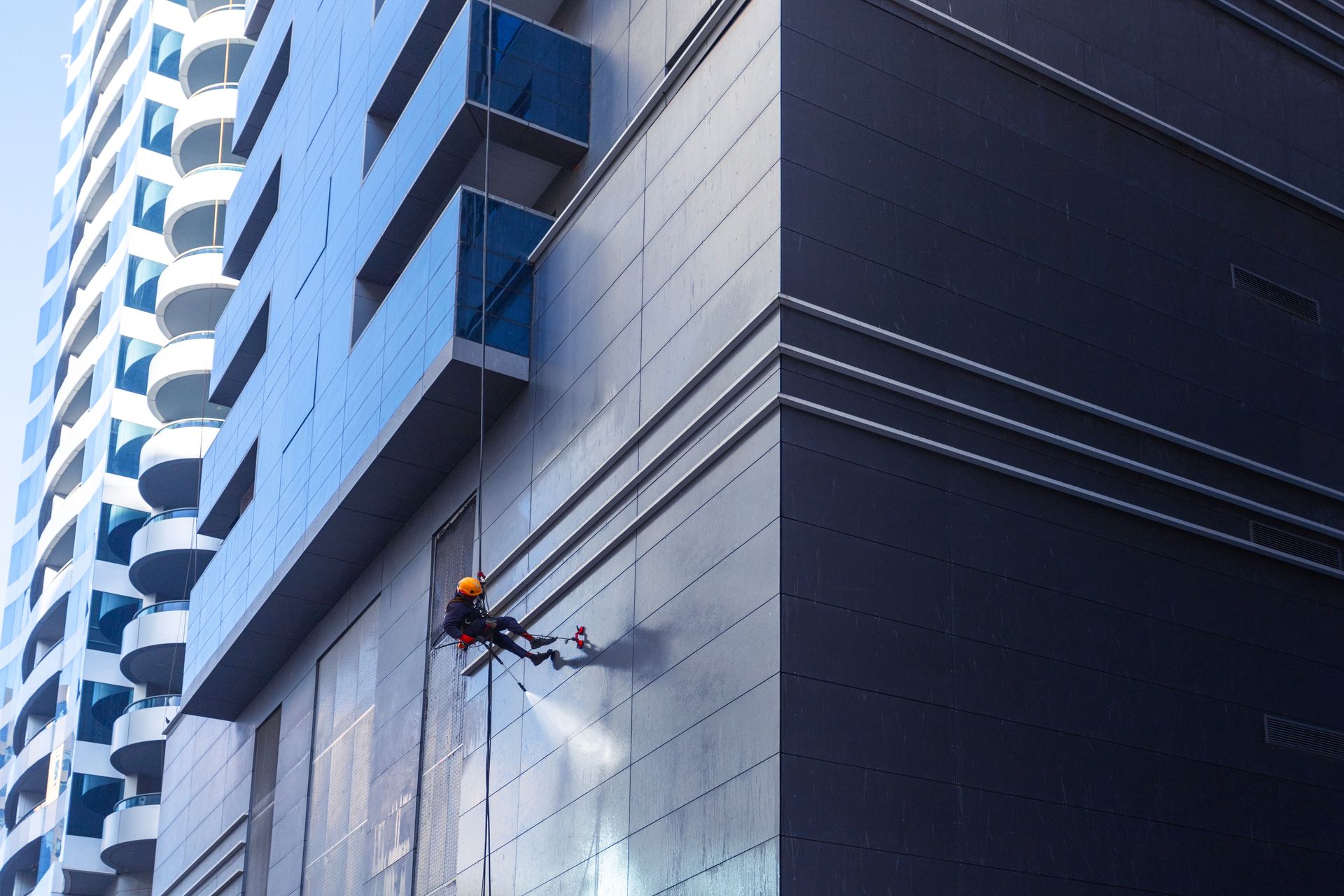 A worker in safety gear rappels down a modern building's exterior, power washing the smooth gray panels.