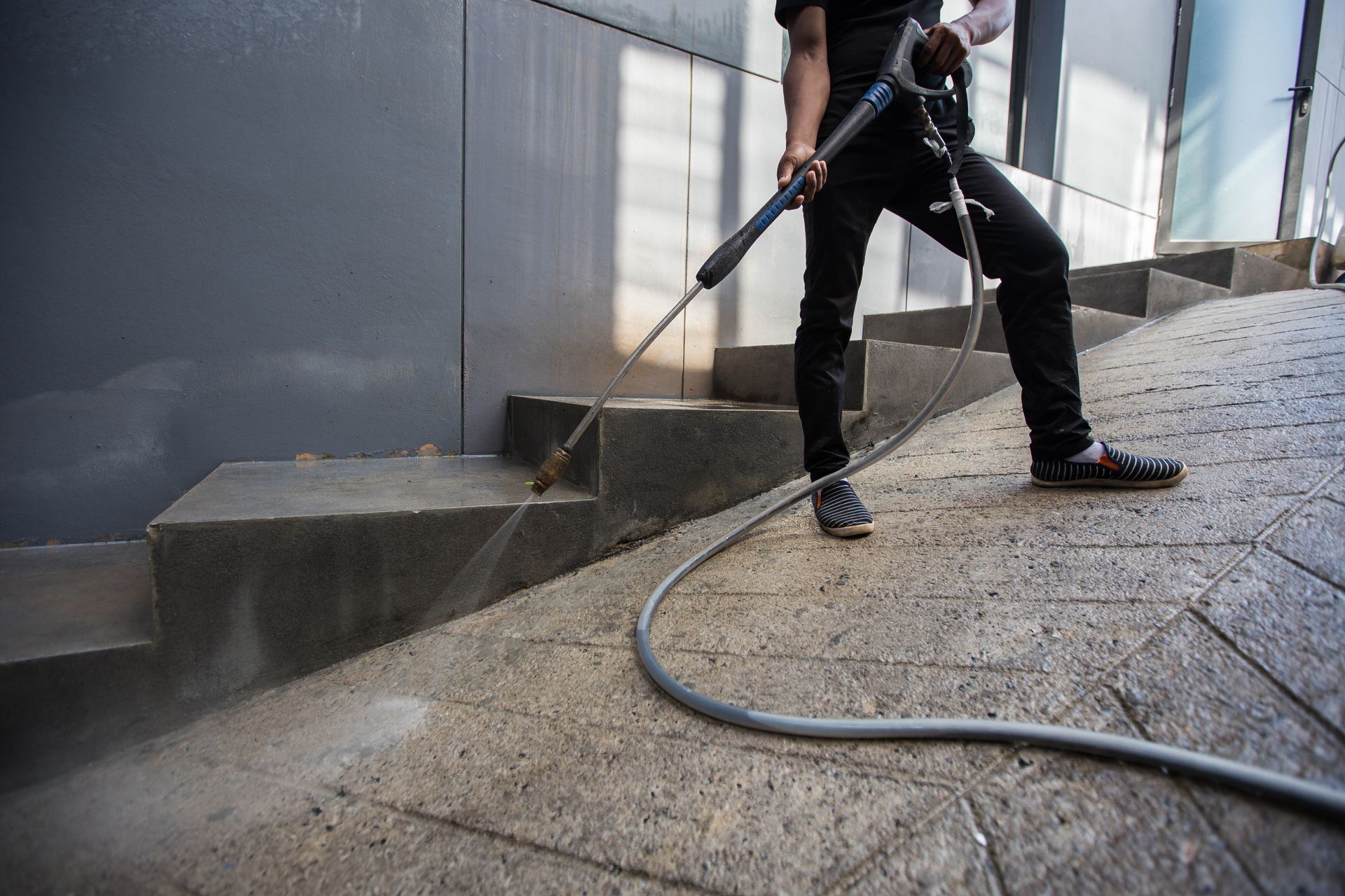 A person in black clothing power washes the stone steps and sloped walkway of a building exterior.