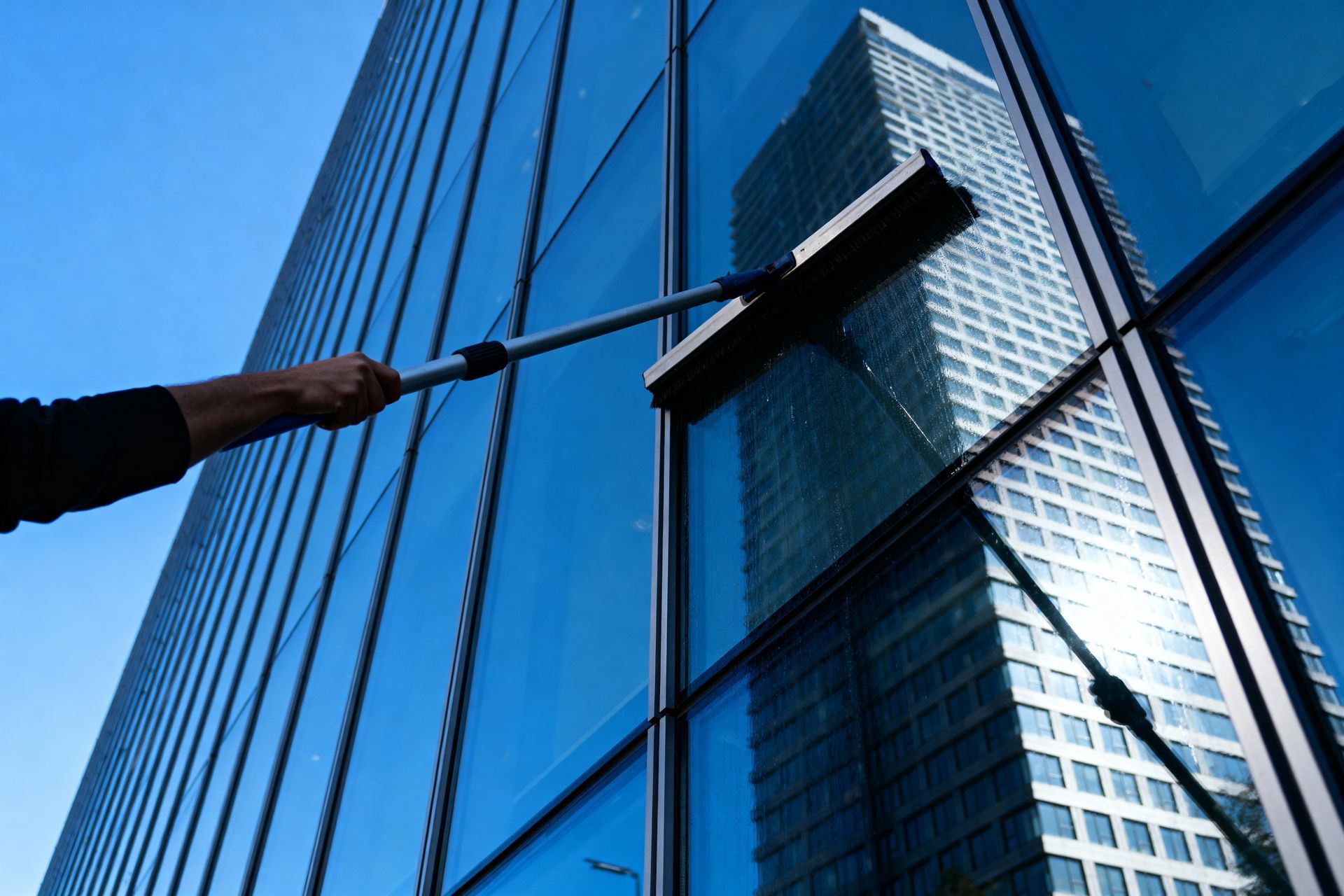 A worker uses an extendable squeegee to clean the glass facade of a skyscraper, which reflects a nearby building.