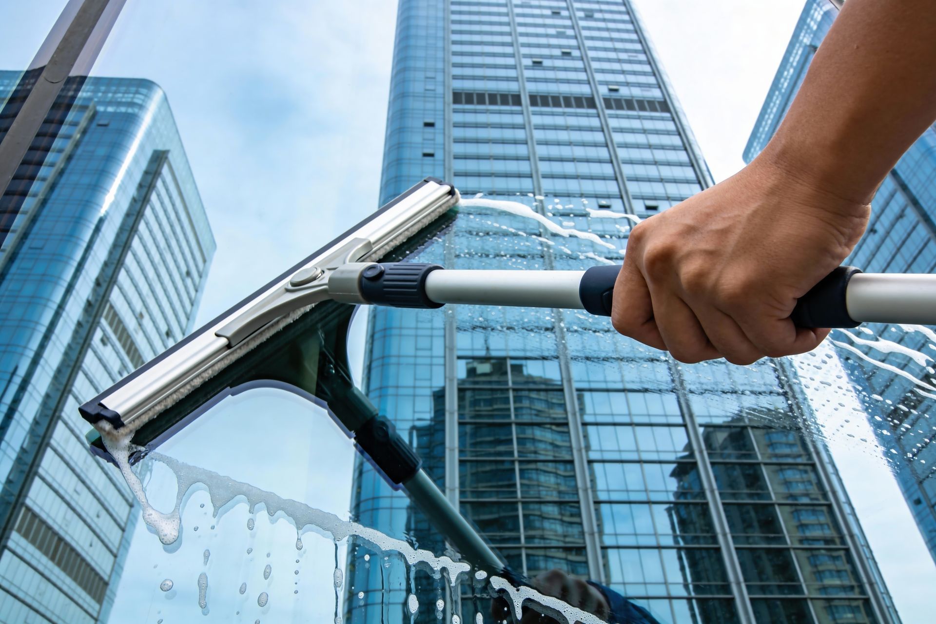 A person’s hand uses a squeegee to clean a soapy glass window on a tall modern skyscraper.